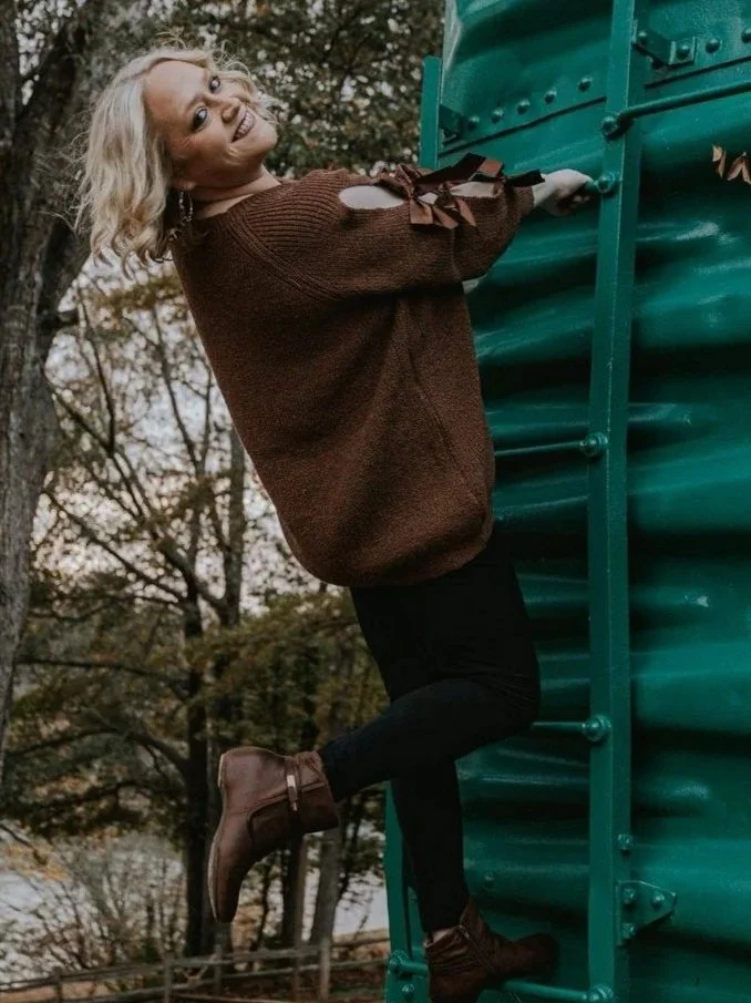 A woman with blonde hair climbing a green metal structure outdoors, wearing a brown sweater, black pants, and brown boots, with trees and autumn foliage in the background.