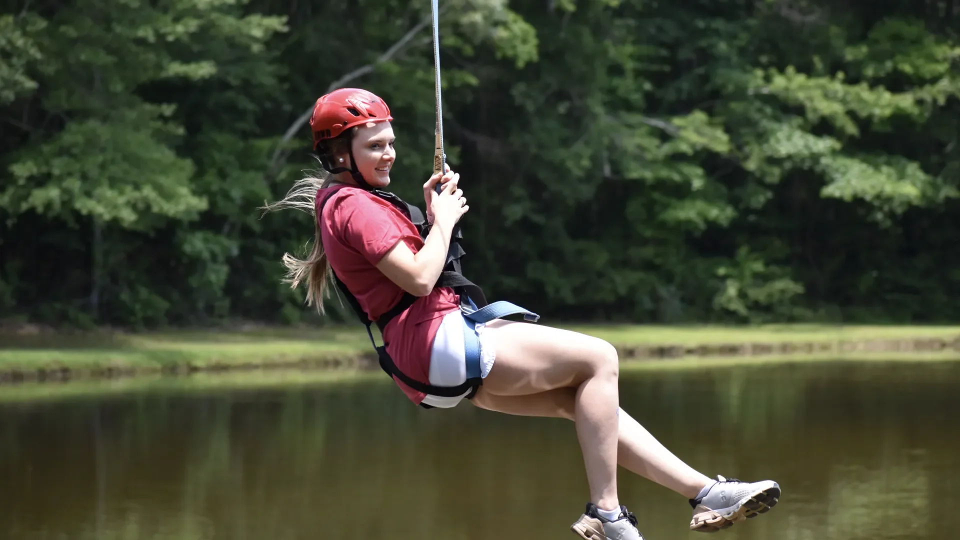 Young woman in red helmet and harness swinging over a body of water in a forested area.