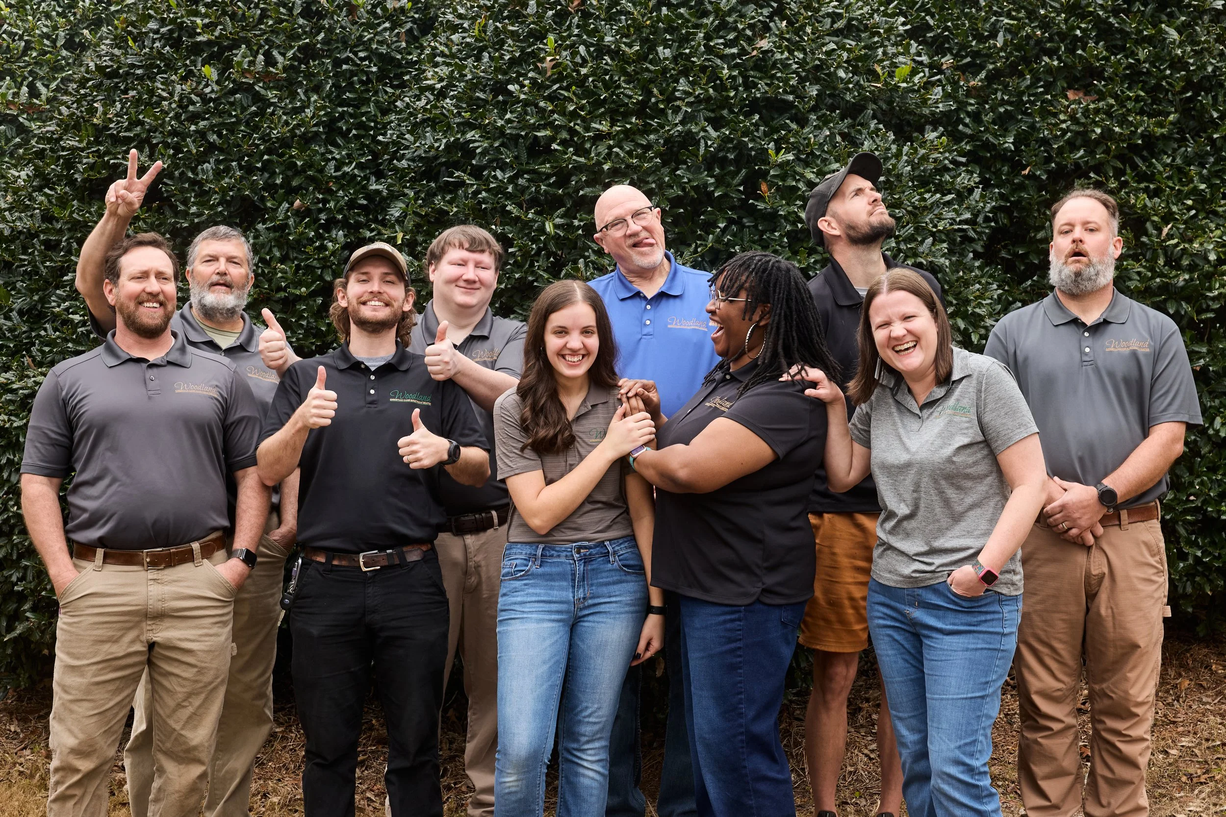 Group of people standing outdoors in front of a hedge, smiling, making gestures like thumbs-up and peace signs, dressed in casual and work clothes.