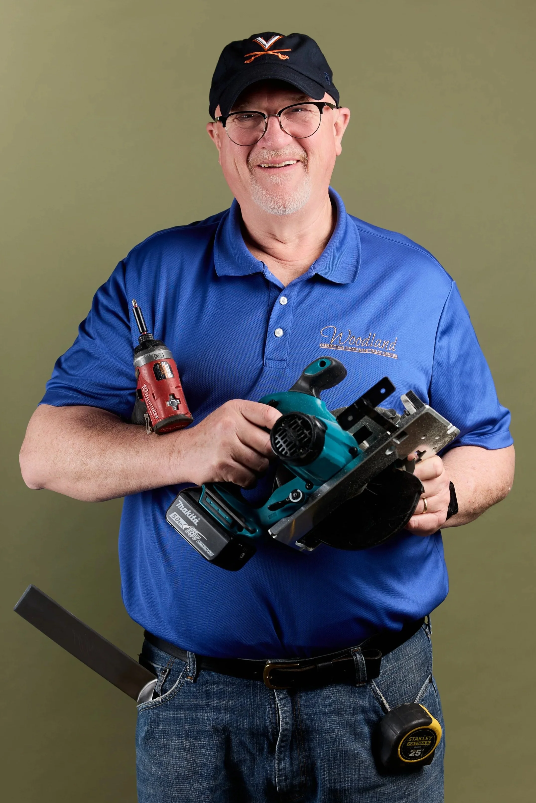 Smiling man wearing glasses, a black cap with Virginia Cavaliers logo, and a blue polo shirt with Woodland logo, holding a cordless power saw and other tools in front of a plain green background.