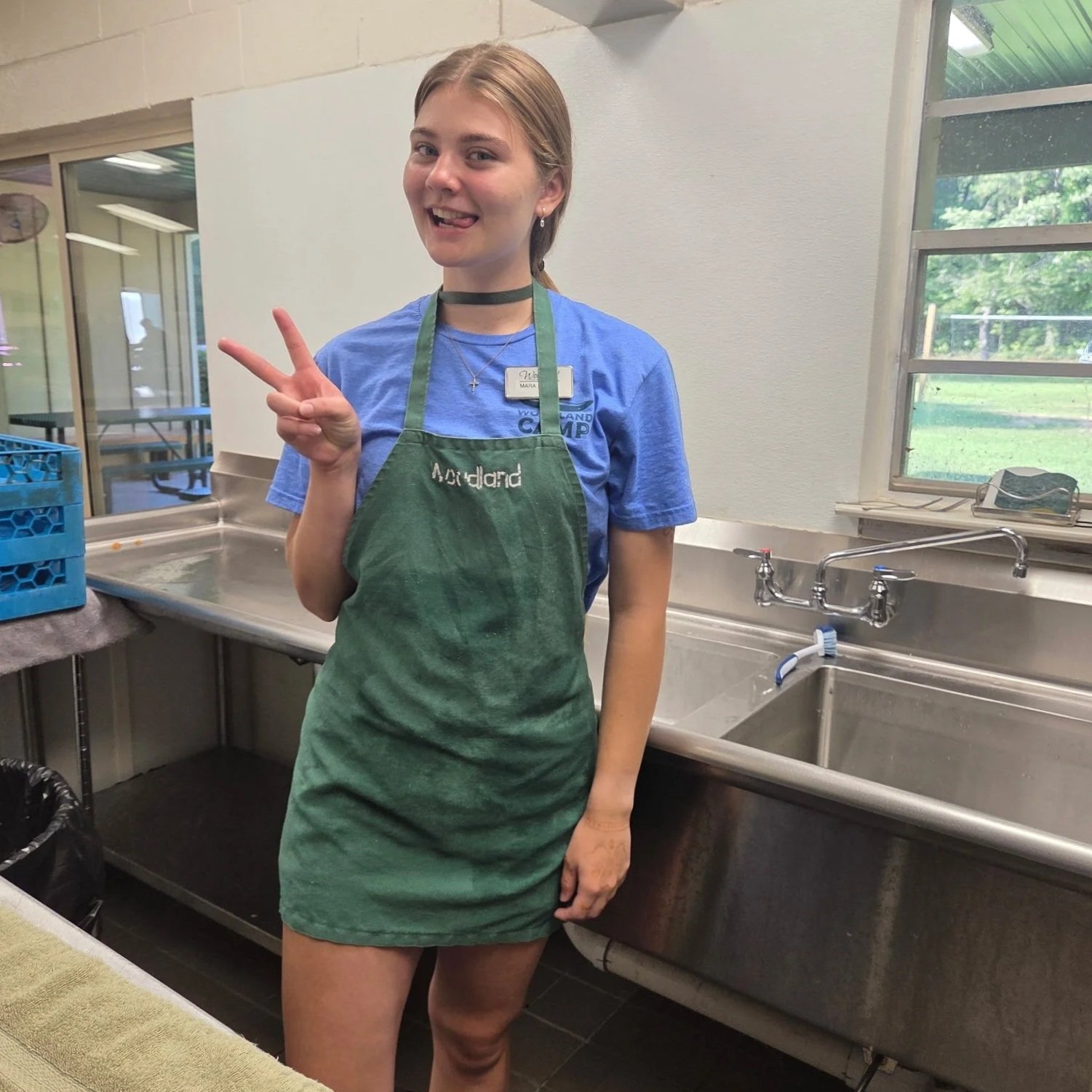 A young woman with blonde hair smiling and making a peace sign with her right hand, wearing a green apron with the word 'Medland' on it, standing in a kitchen by a large stainless steel sink with a window behind her showing a grassy outdoor area.