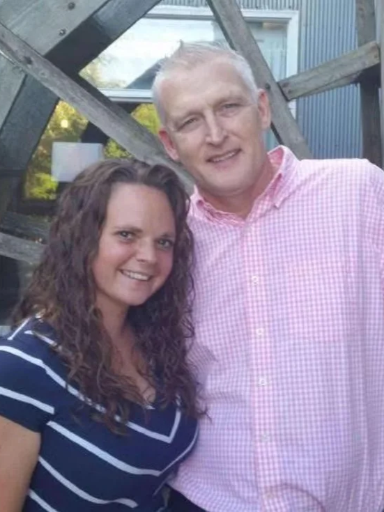 A woman with long, curly brown hair and a man with short, light blond hair smiling and posing outdoors in front of a wooden structure and a house.