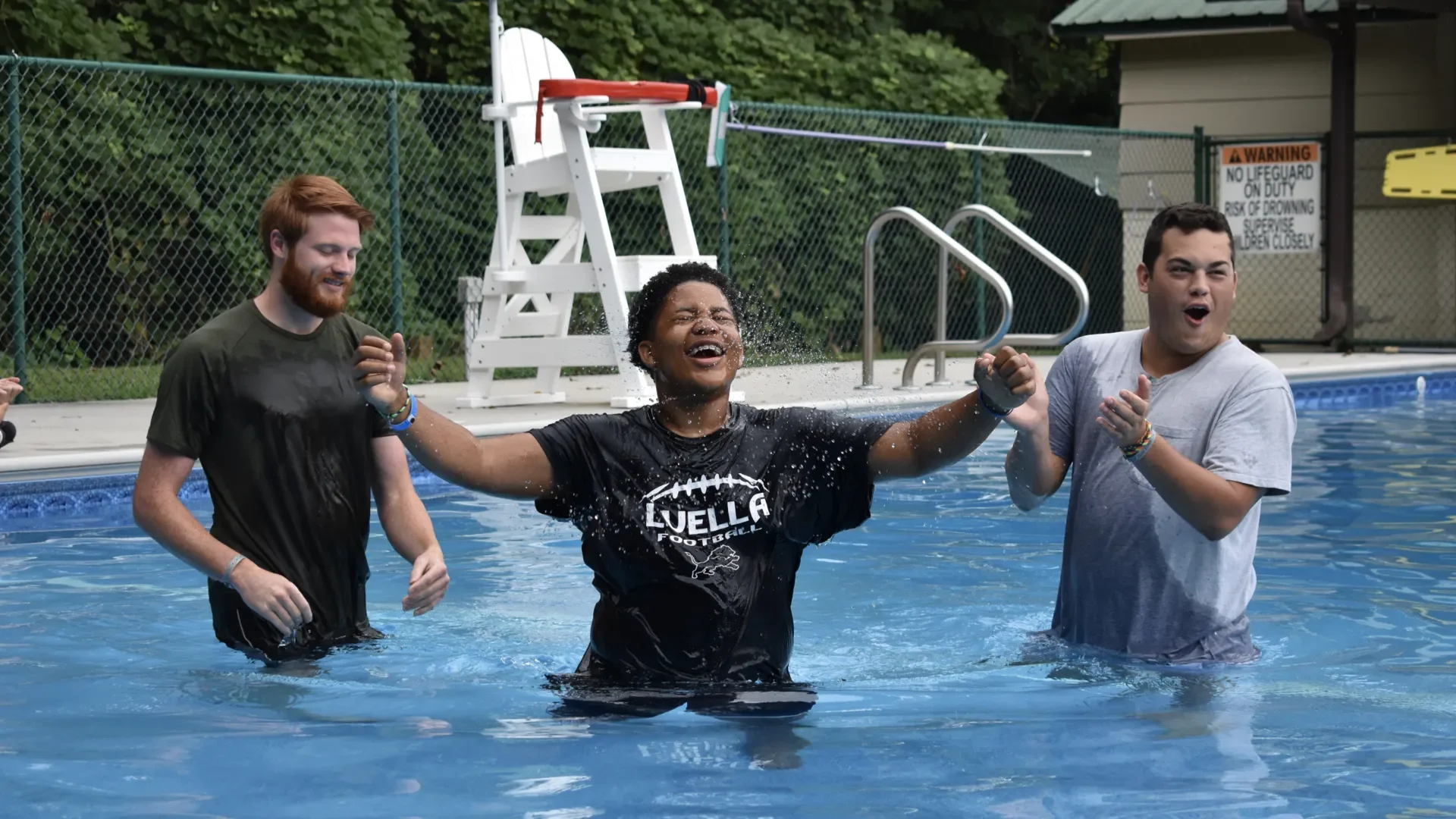 Three young people in a swimming pool, celebrating and smiling, with one person in the middle raising their arms, wearing a black football T-shirt, while the other two, wearing gray shirts, stand on the sides.