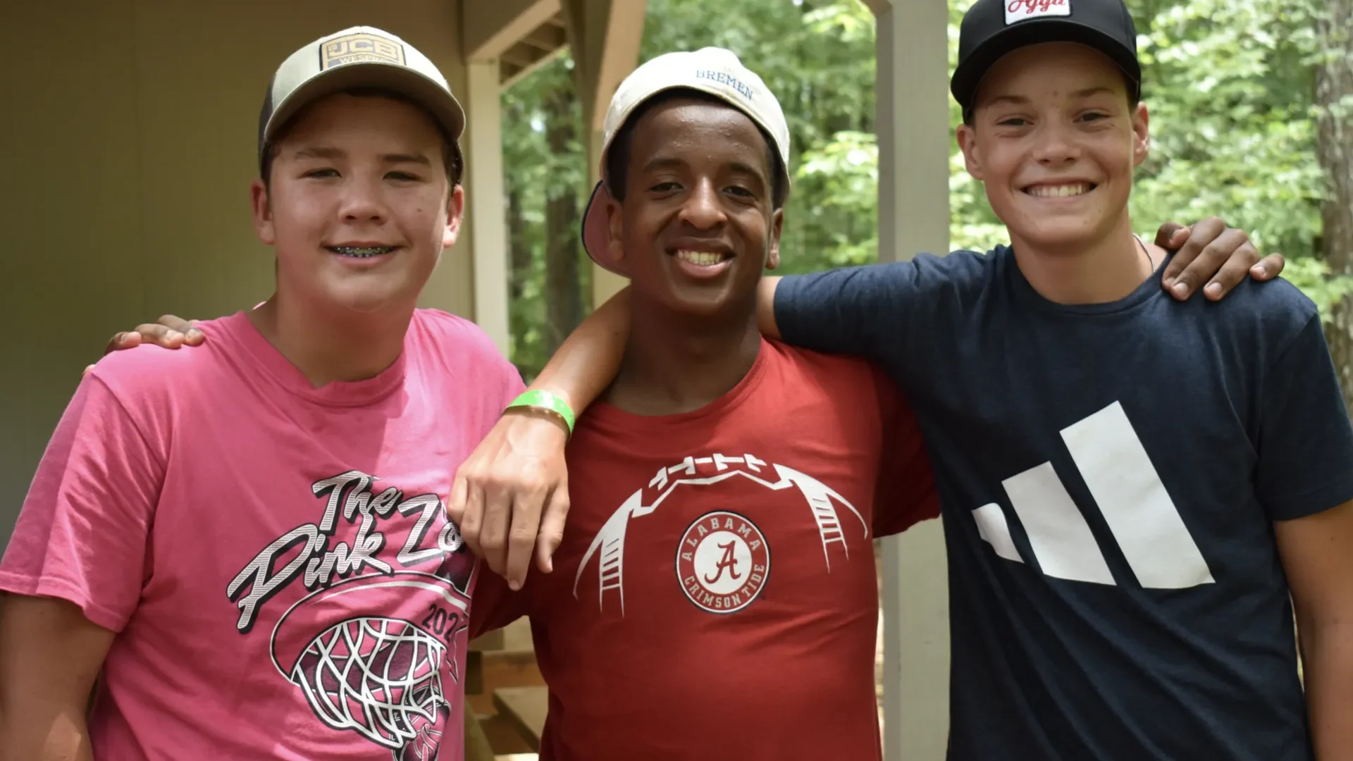 Three smiling boys with their arms around each other outdoors, standing in front of trees and a beige building. The boy on the left is wearing a pink T-shirt and a beige cap, the boy in the middle is wearing a red T-shirt with a football logo and a w
