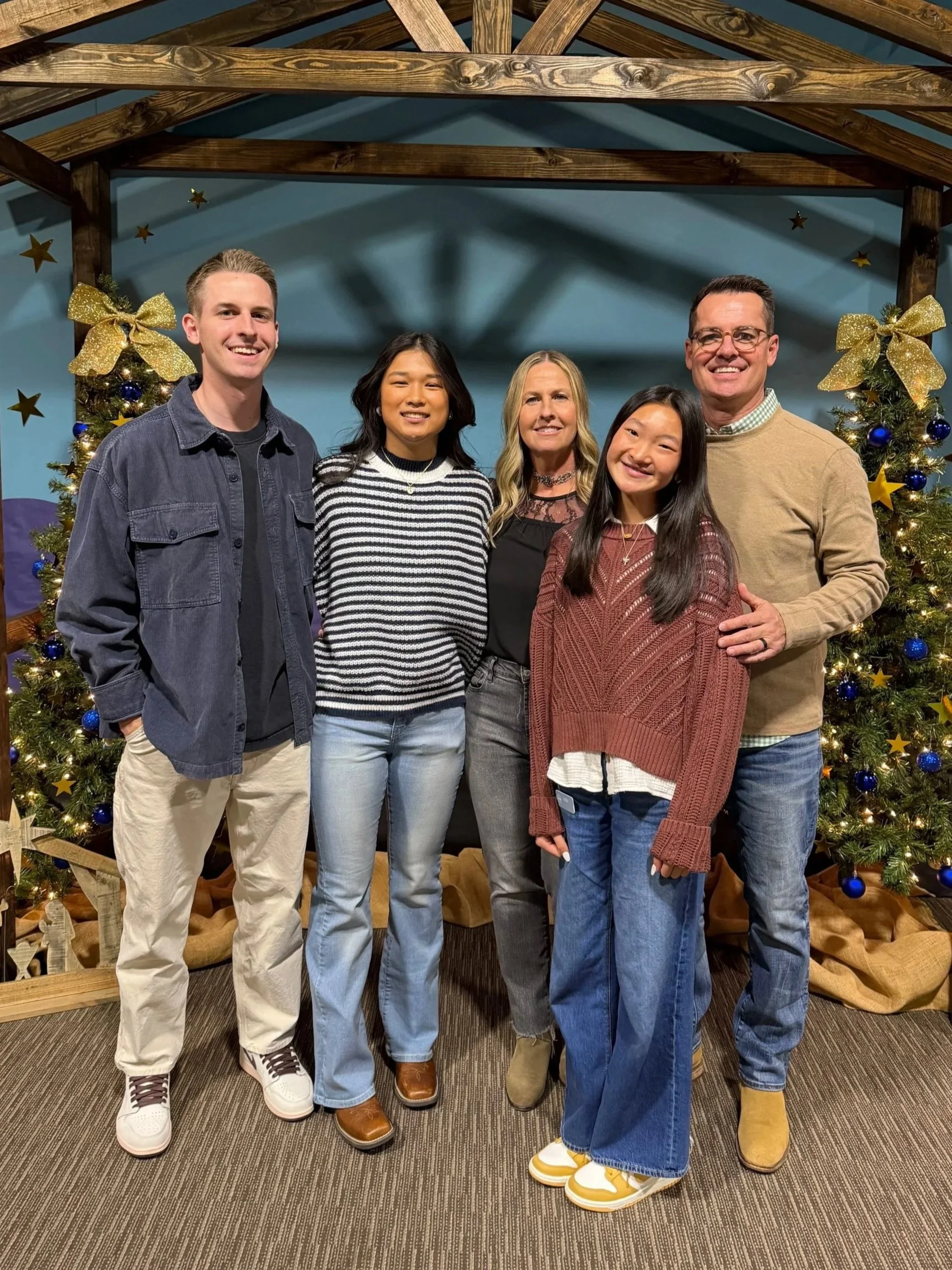 A group of five people, three women and two men, standing in front of decorated Christmas trees with gold bows, blue ornaments, and gold stars, indoors with a wooden roof structure in the background.