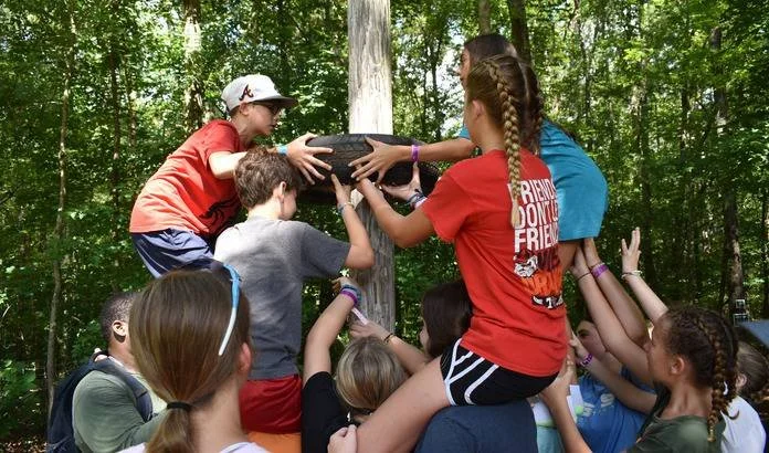 Children working together outdoors during a team-building activity, lifting a large log while standing around a tree in a wooded area.
