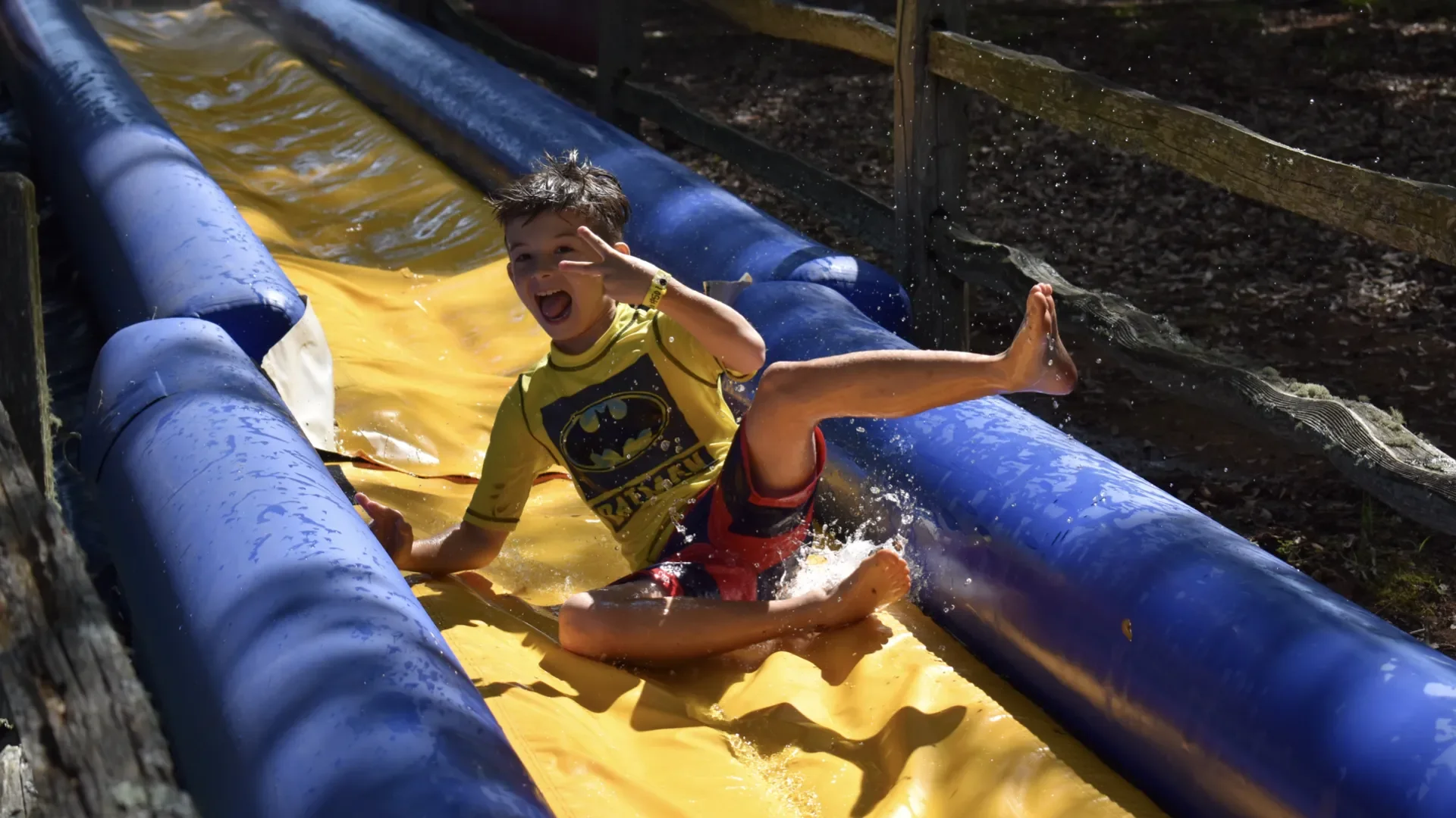 Boy in a yellow Batman T-shirt and red shorts sliding down a yellow water slide with blue sides, smiling and making a peace sign with hand.
