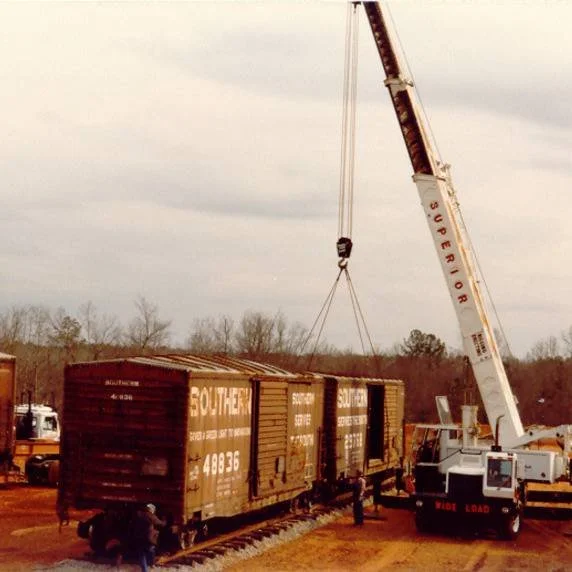 A large crane labeled 'Super B' lifts a freight train car during transport or construction. Several people are nearby, and the scene is set outdoors with a cloudy sky.