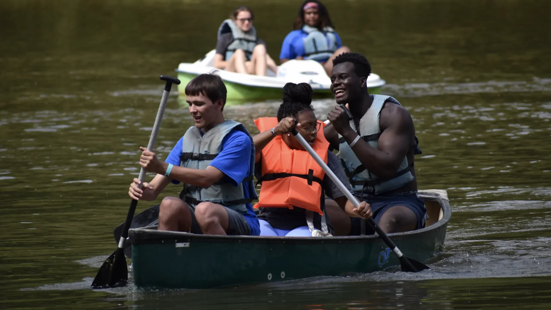 Four people are paddling a canoe on a lake, two in the front and two in the back, with two more people in a small boat in the background.
