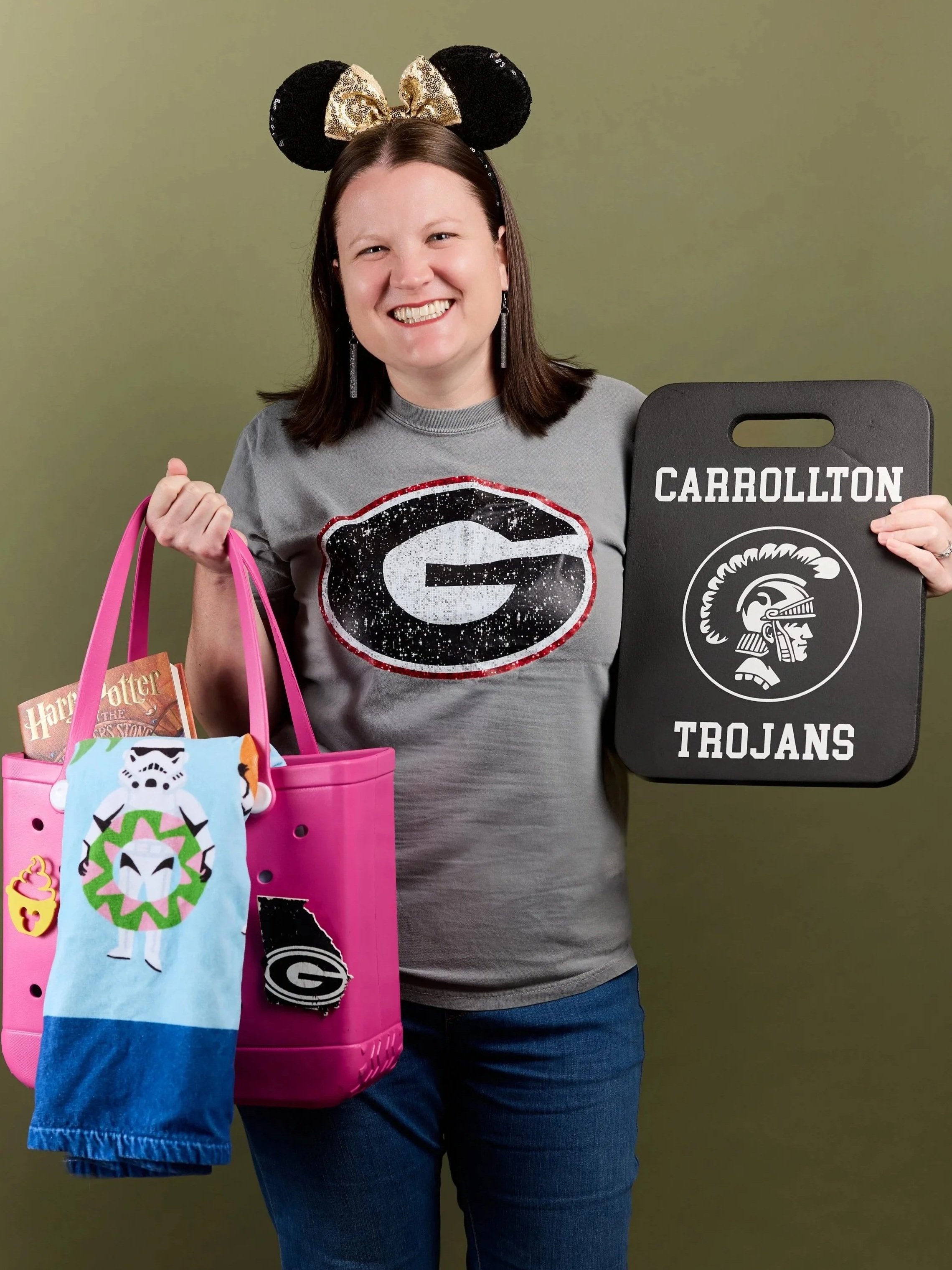 Woman smiling, wearing Georgia Bulldogs headband and t-shirt, holding pink tote bag with Harry Potter book and sock with Star Wars stormtrooper and Georgia G logo, and a black tray with 'Carrollton Trojans' logo and name.
