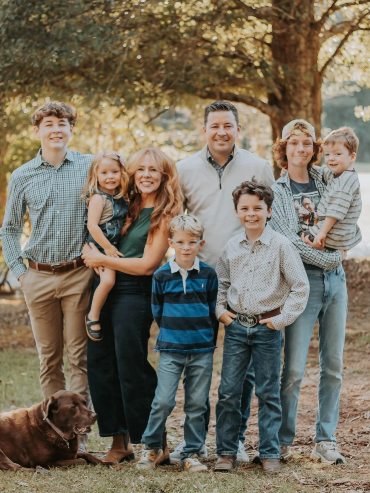 Family portrait outdoors featuring a group of adults and children, with a dog on the ground, under a large tree.