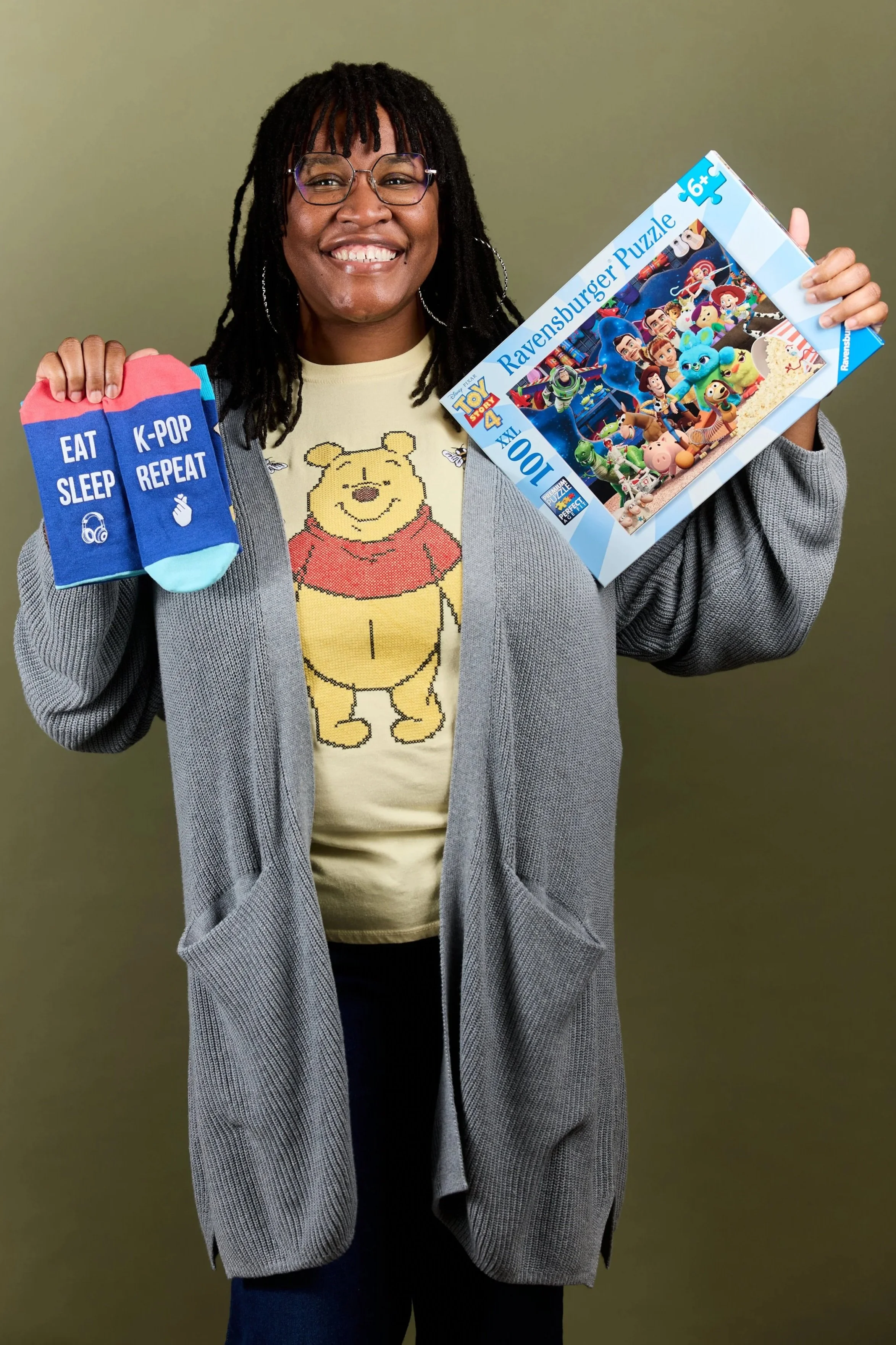 A smiling person with glasses and dreadlocks holding a Toy Story 4 Ravensburger puzzle and a pair of colorful socks with the words "Eat Sleep K-Pop Repeat".