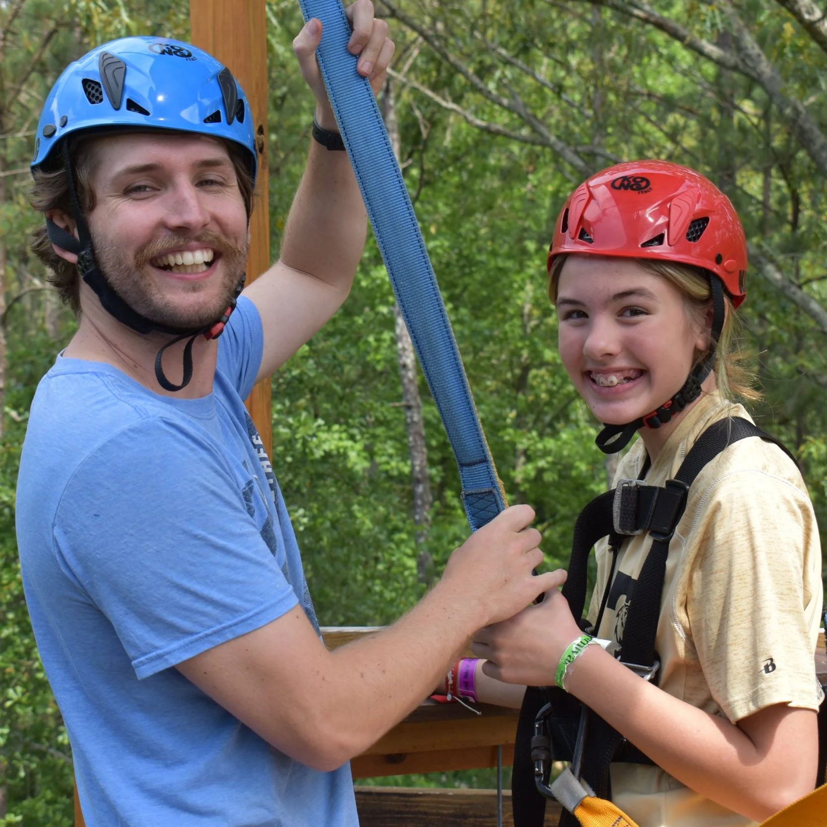 A man and girl, both wearing helmets and harnesses, smiling and holding a blue strap outdoors in a wooded area.