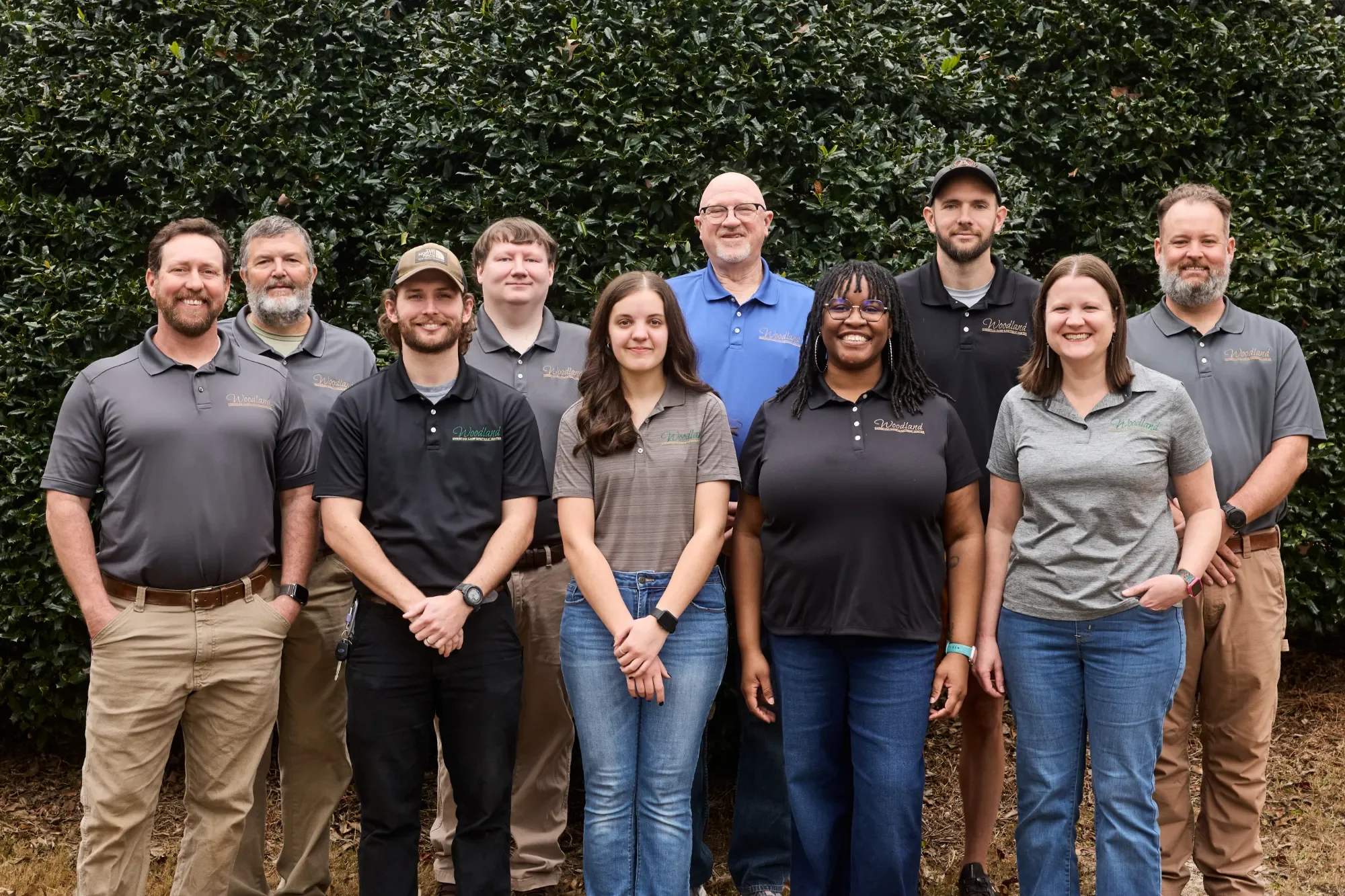 A group of ten people standing outdoors in front of leafy bushes, posing for a photo. They are dressed in casual and company-branded shirts.