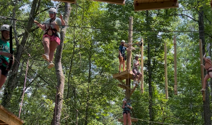 Children participating in a high ropes course adventure in a wooded area with ropes, platforms, and safety harnesses.