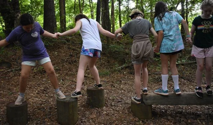 Five children holding hands while walking on a balance beam in a forested area.