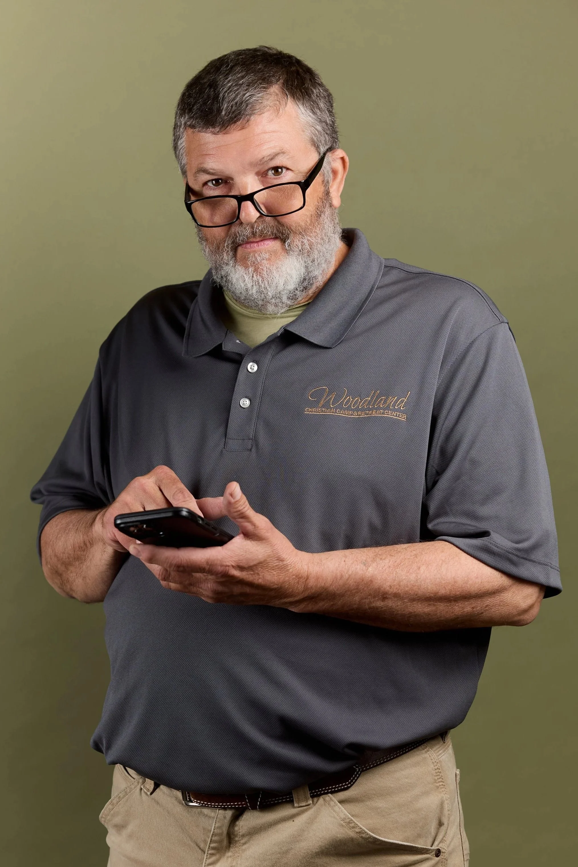 A middle-aged man with gray hair, beard, and glasses, wearing a gray polo shirt with «Woodland» embroidered on it, standing against a green background and looking at his phone.
