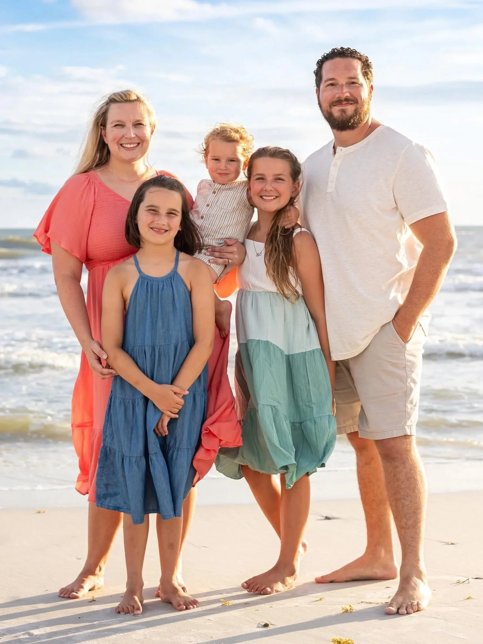 A family of six posing on the beach. The group includes two adults and four children, all smiling, with the ocean and sky in the background.