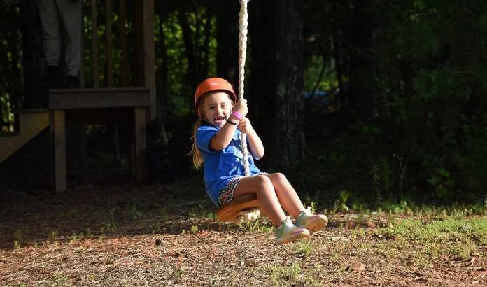 A young girl with a helmet playing on a zip line in a wooded outdoor area.