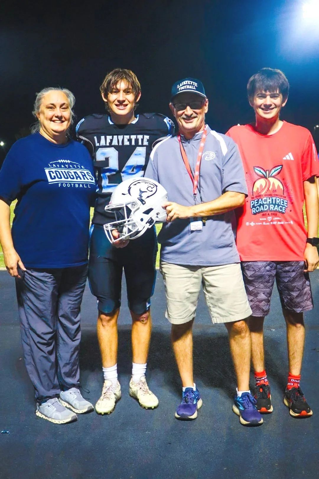 A woman, a football player, a coach, and a young man stand together on a football field at night, smiling. The woman and the young man wear Lafayette football shirts, while the football player holds a helmet.
