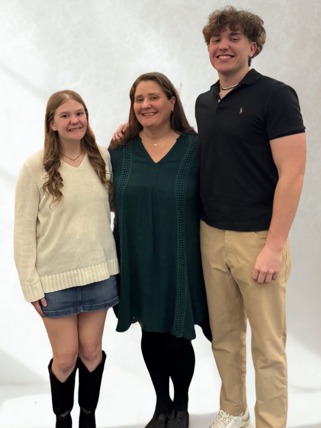 A woman and two teenagers, a girl and a boy, standing together smiling for the camera in a plain indoor setting.