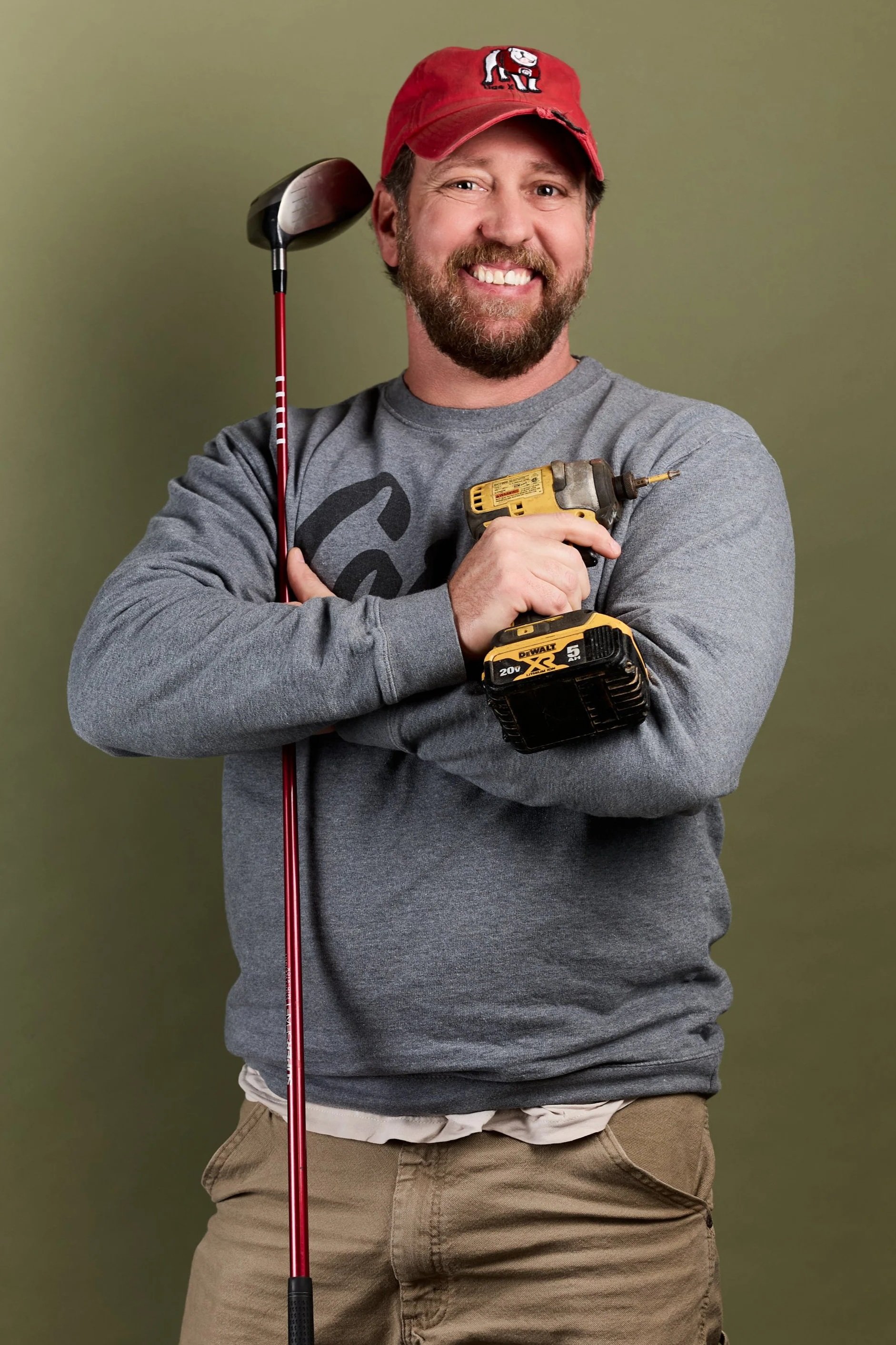 A man with a beard and mustache wearing a red cap with a white emblem, a gray sweatshirt, and khaki pants. He is smiling, holding a yellow cordless drill, with a golf club resting on his shoulder.