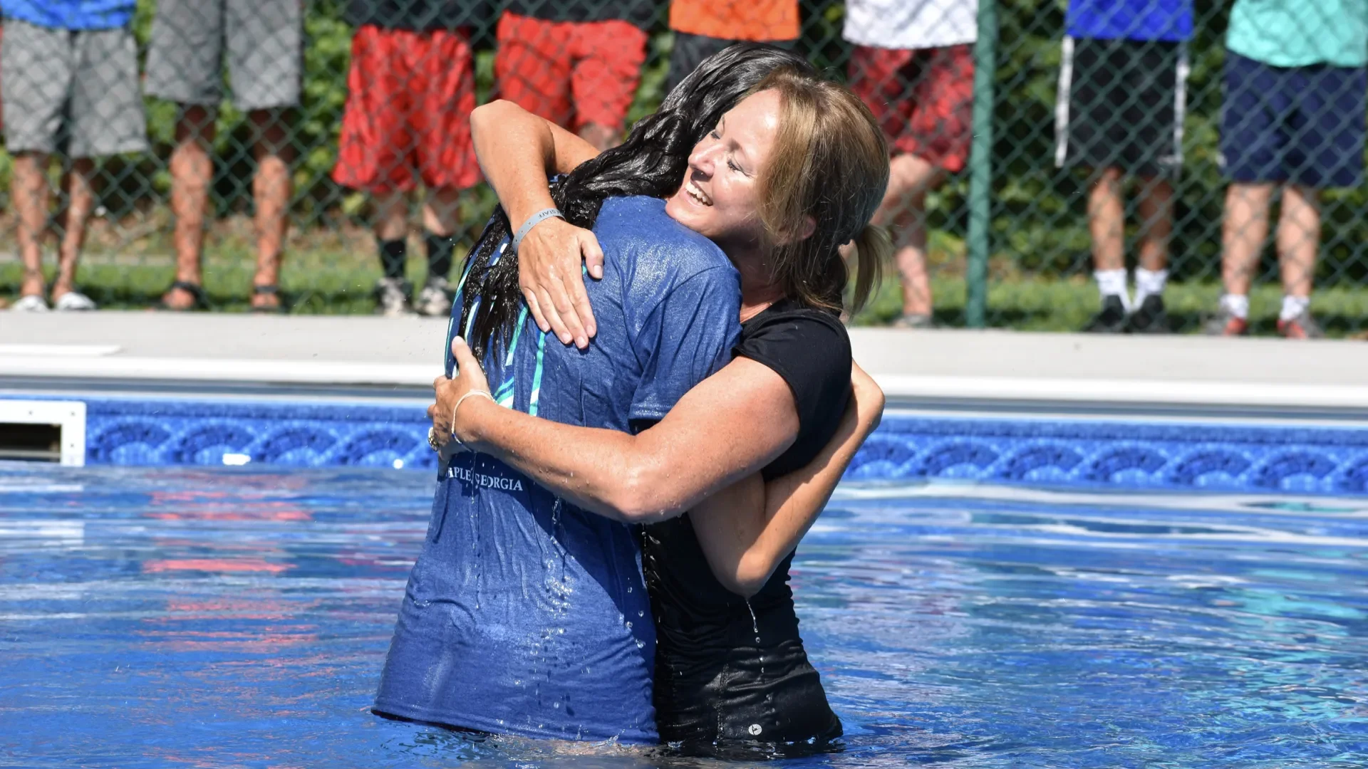 Two women hugging in a swimming pool with a chain-link fence and children in swim trunks in the background.
