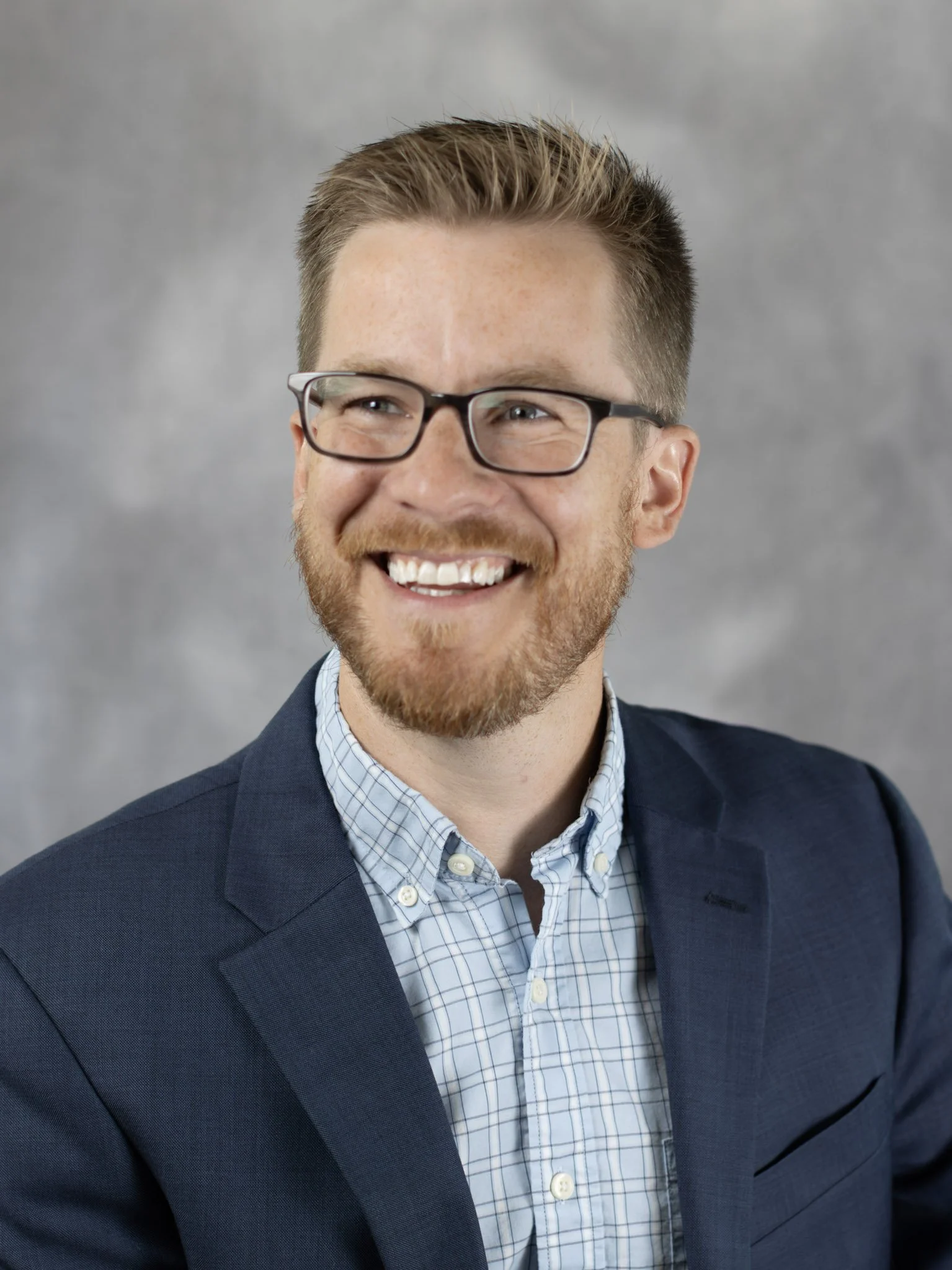 A man with blonde hair, glasses, and a beard wearing a blue suit jacket and light blue shirt smiling against a gray background.