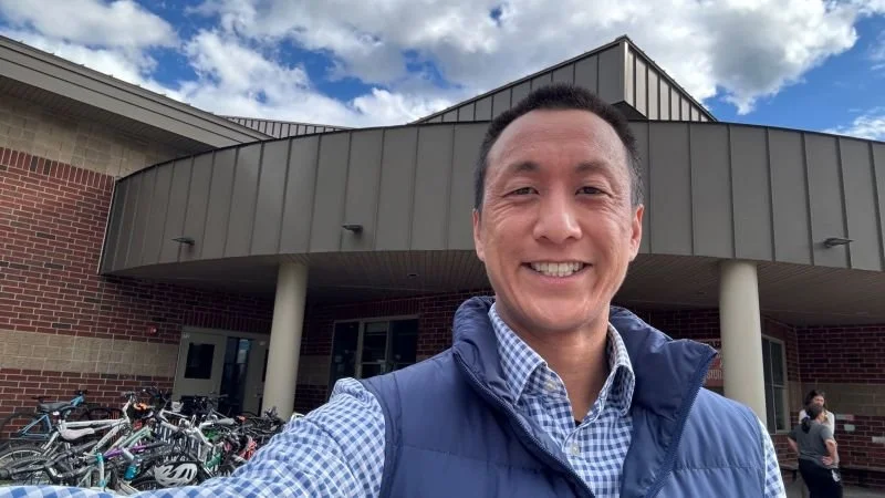 A smiling man taking a selfie outside a brick building with a covered entrance, bicycles parked nearby, and a partly cloudy sky overhead.