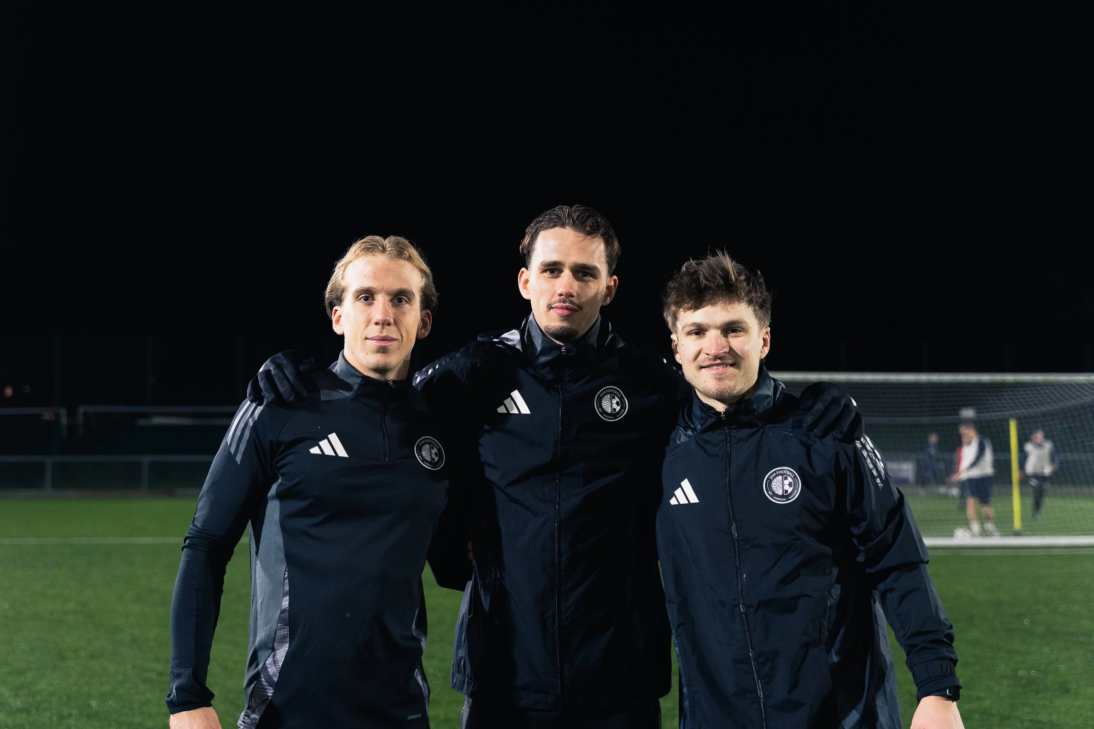 Three young men in sports jackets with team logos, standing close together on a soccer field at night, smiling, with a goal in the background.