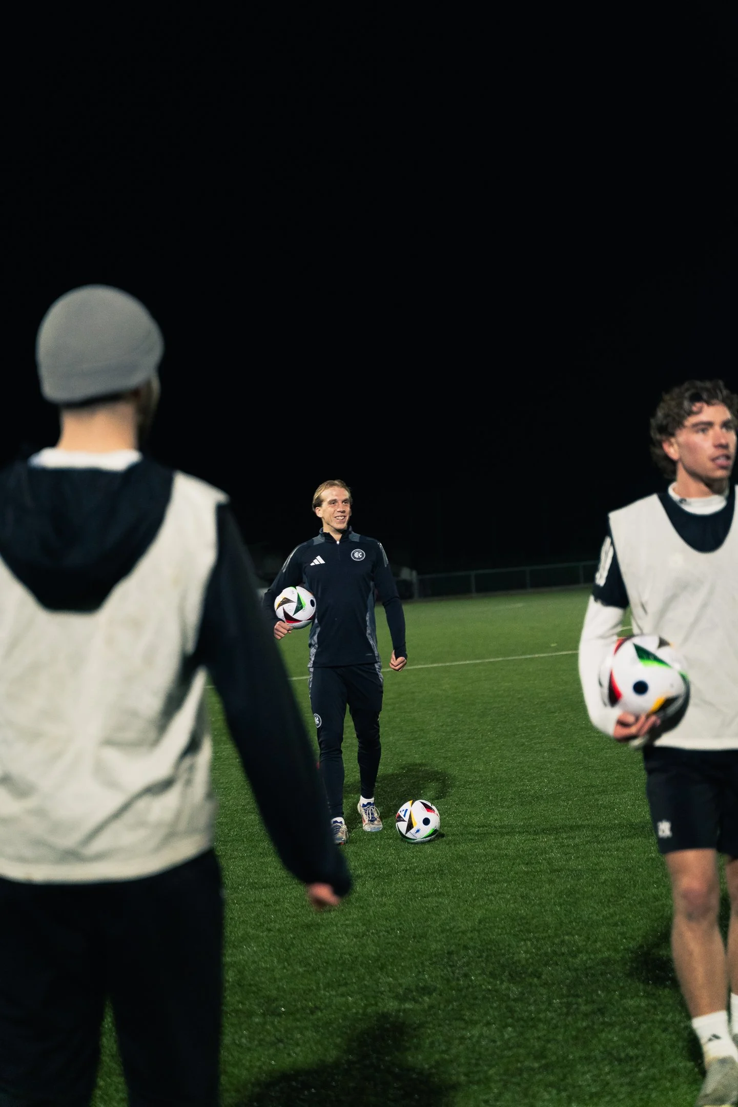 Soccer practice at night with a coach and two players on the field, holding soccer balls.