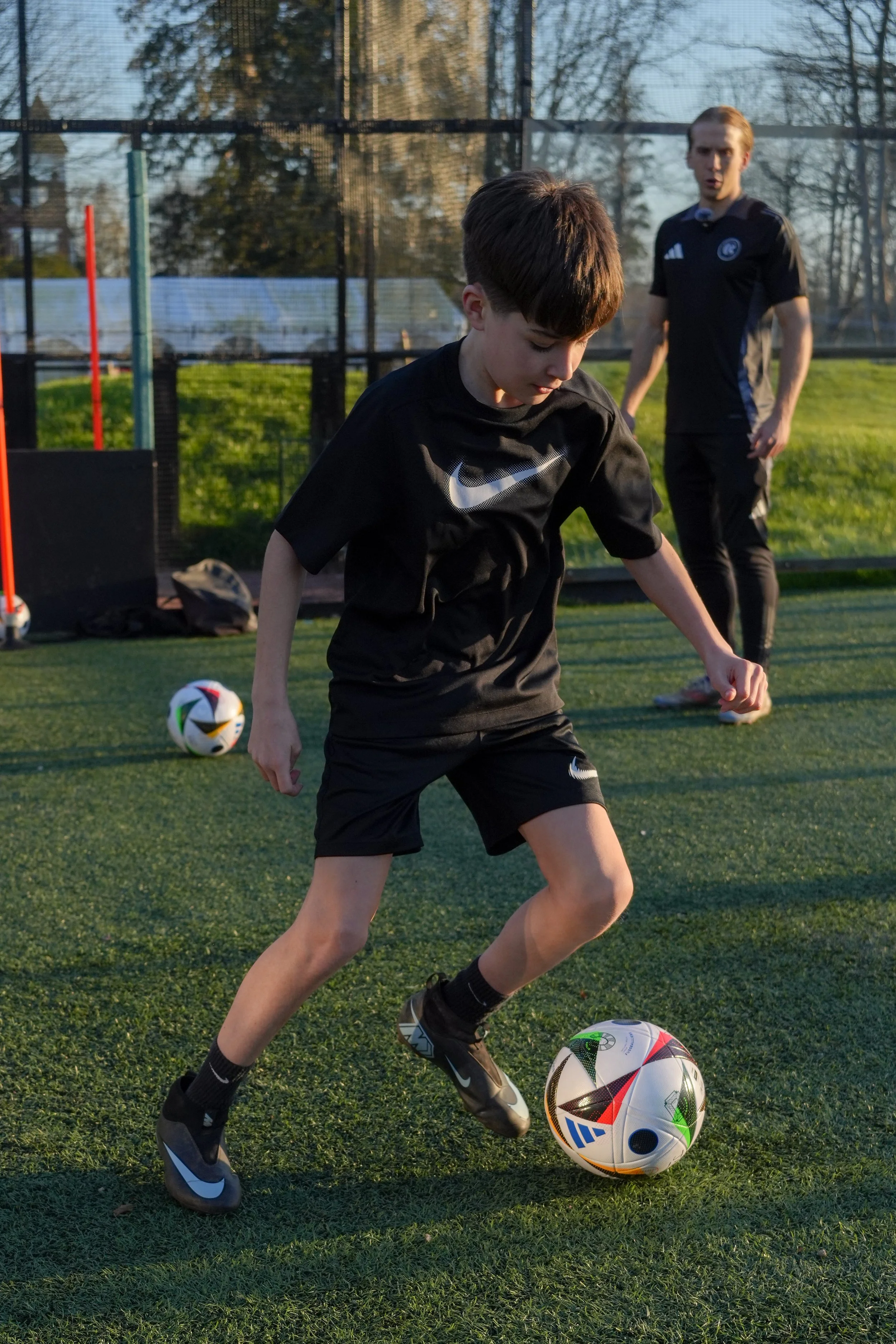 A young boy in black athletic clothing dribbling a soccer ball on a green field, with an adult woman in the background watching.