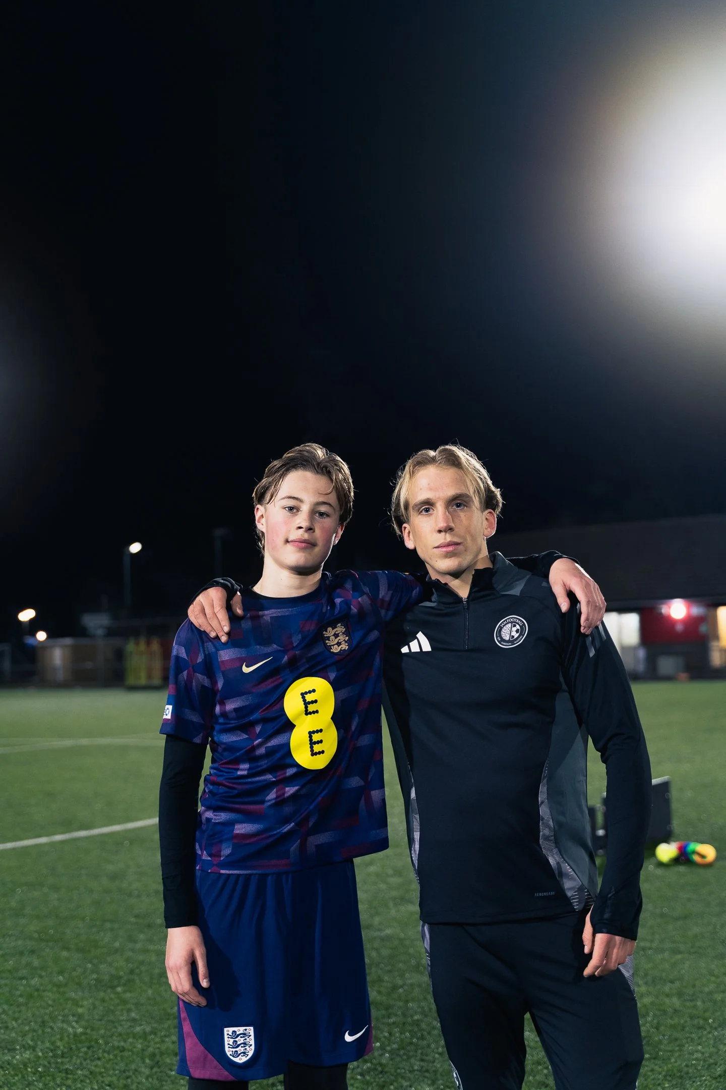 Two young males standing on a soccer field at night, with arms around each other's shoulders, wearing athletic gear.