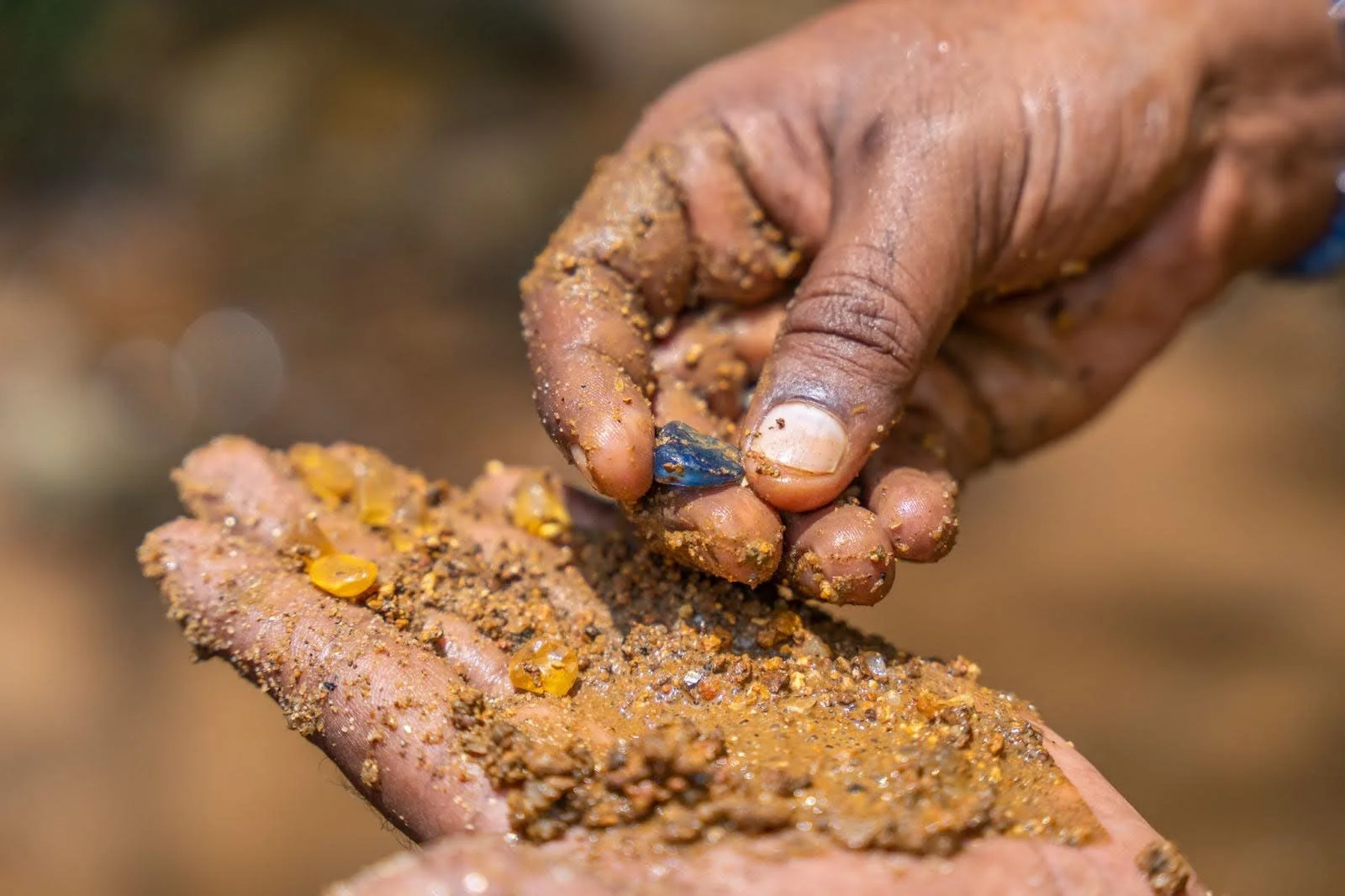 Close-up of a person's hand picking up a shiny blue gemstone from a pile of sand and small rocks.