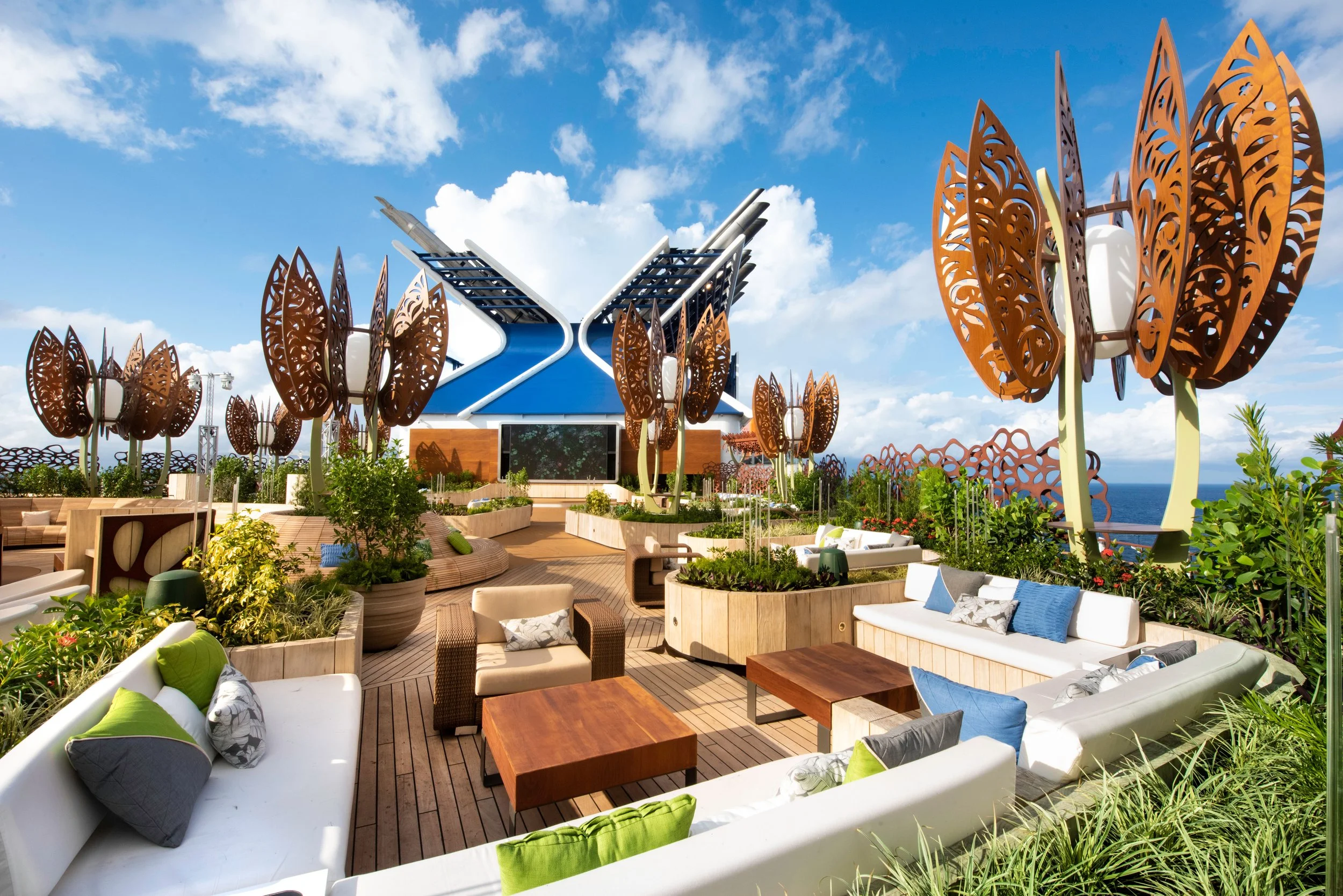 A lush, green Rooftop Garden on a Celebrity Edge-class ship featuring whimsical metal tree sculptures, cozy seating nooks, and a large outdoor movie screen.