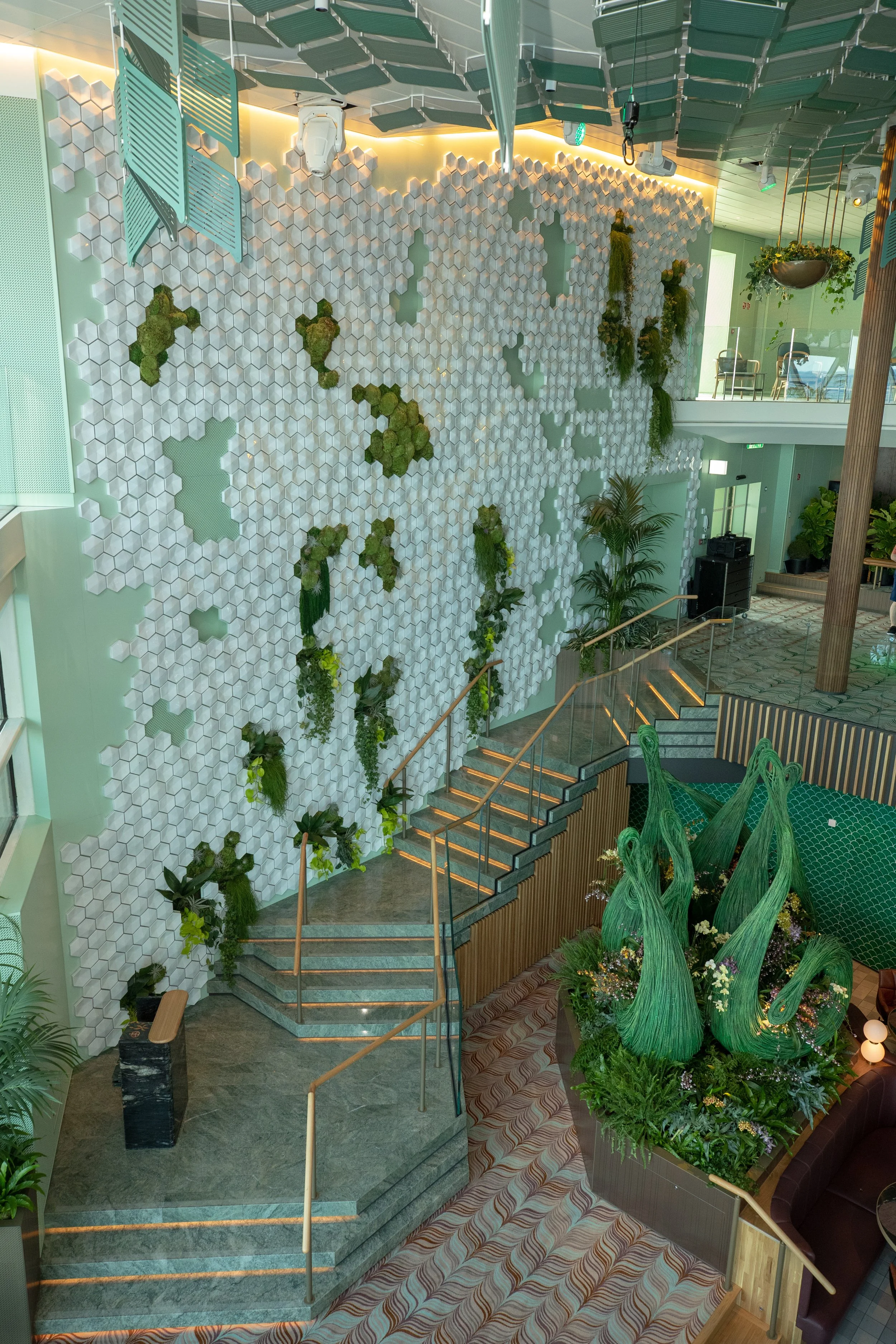 The multi-level Eden lounge on a Celebrity Edge-class ship, featuring floor-to-ceiling glass windows, lush winding greenery, and modern architectural seating overlooking the ocean.