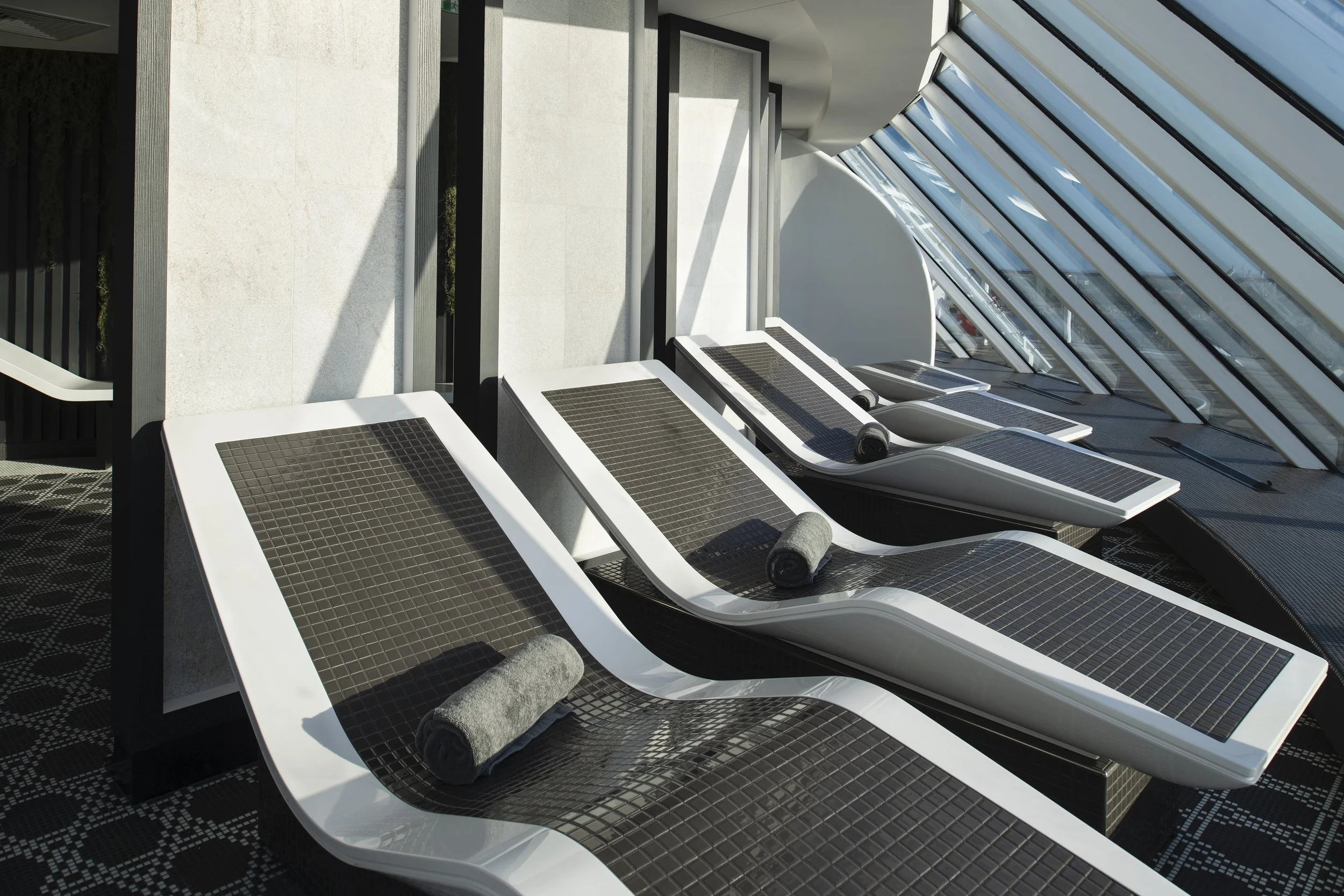 Ergonomically designed stone lounge chairs with a heated surface, tiled in mosaic patterns and facing floor-to-ceiling windows with a view of the ocean in the SEA Thermal Suite.