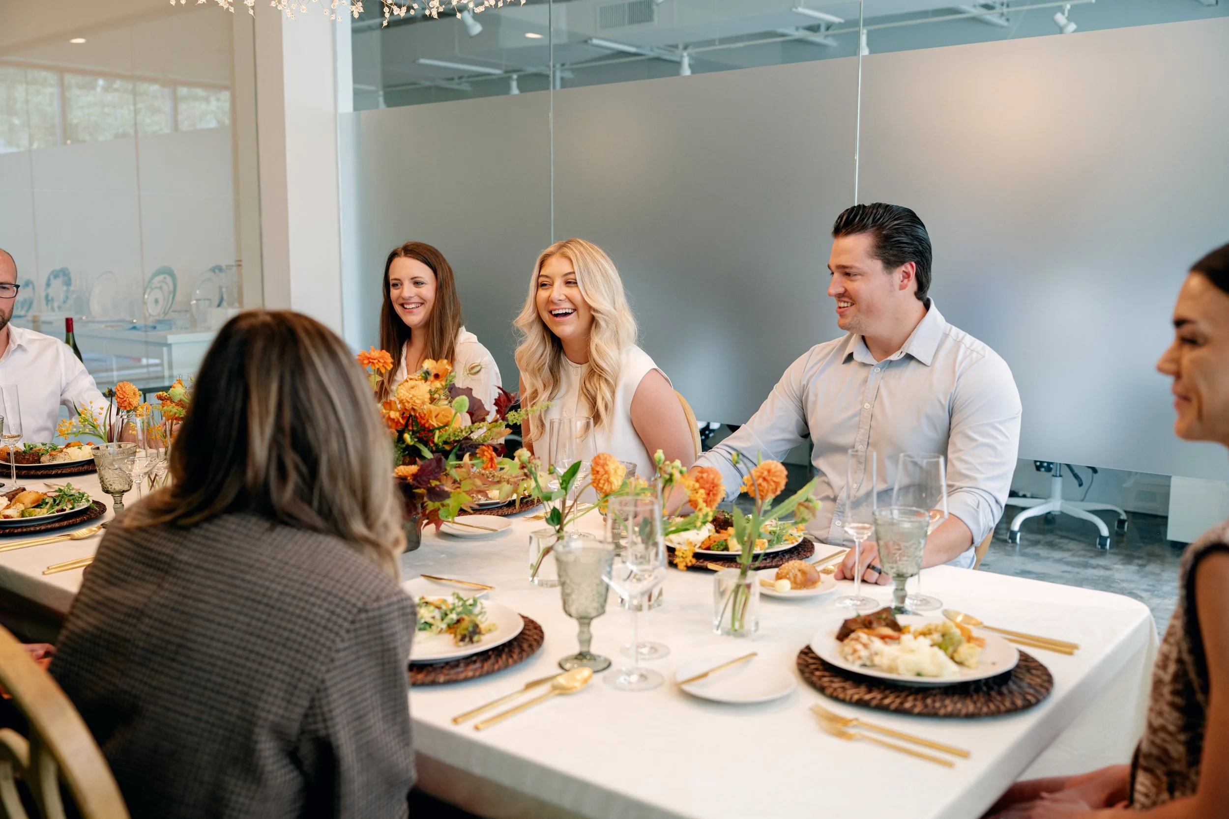 People gathered around a dining table with floral centerpieces, enjoying a meal and conversation in a bright, modern room.
