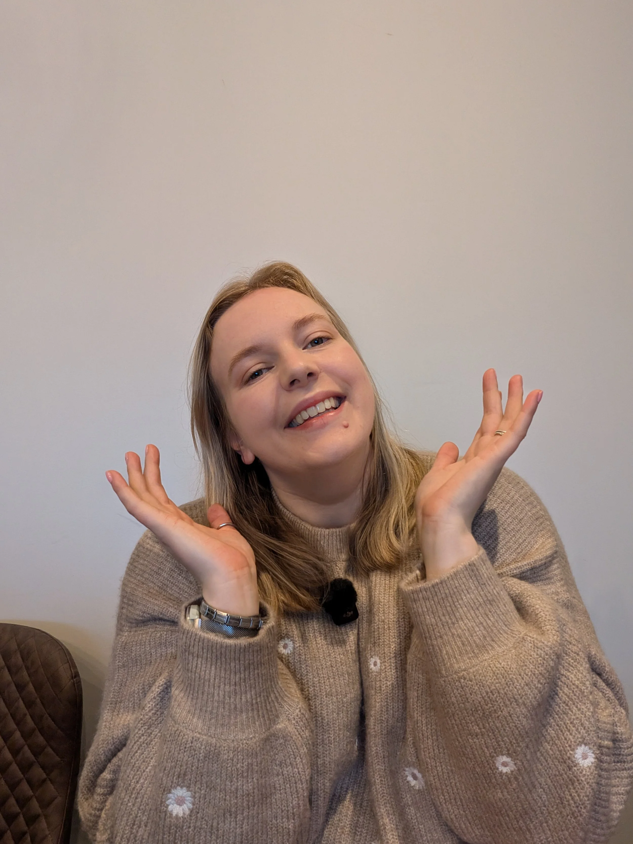 A copywriter with blonde hair is smiling and making playful gestures with her hands. She is wearing a beige sweater with small floral embroidery, a wristwatch, and a ring, sitting against a plain gray wall.