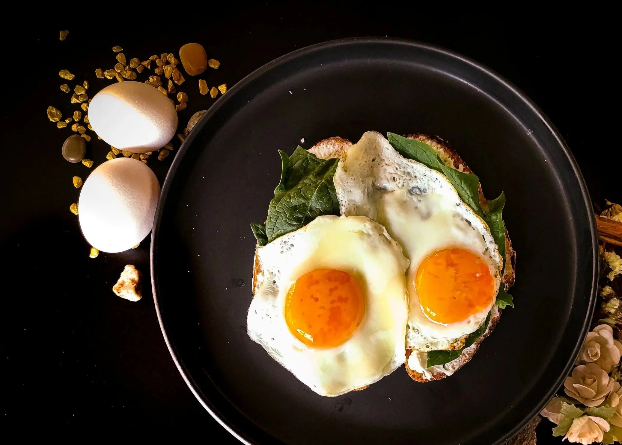 Fried eggs on toast with spinach on a black plate, with two eggs and some small stones and a shell on a dark surface.