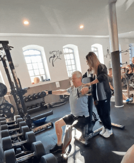 Ein älterer Mann trainiert an einem Kraftgerät im Fitnessstudio, während eine junge Frau ihn unterstützt. Im Hintergrund sind Hanteln und Fitnessgeräte sichtbar.