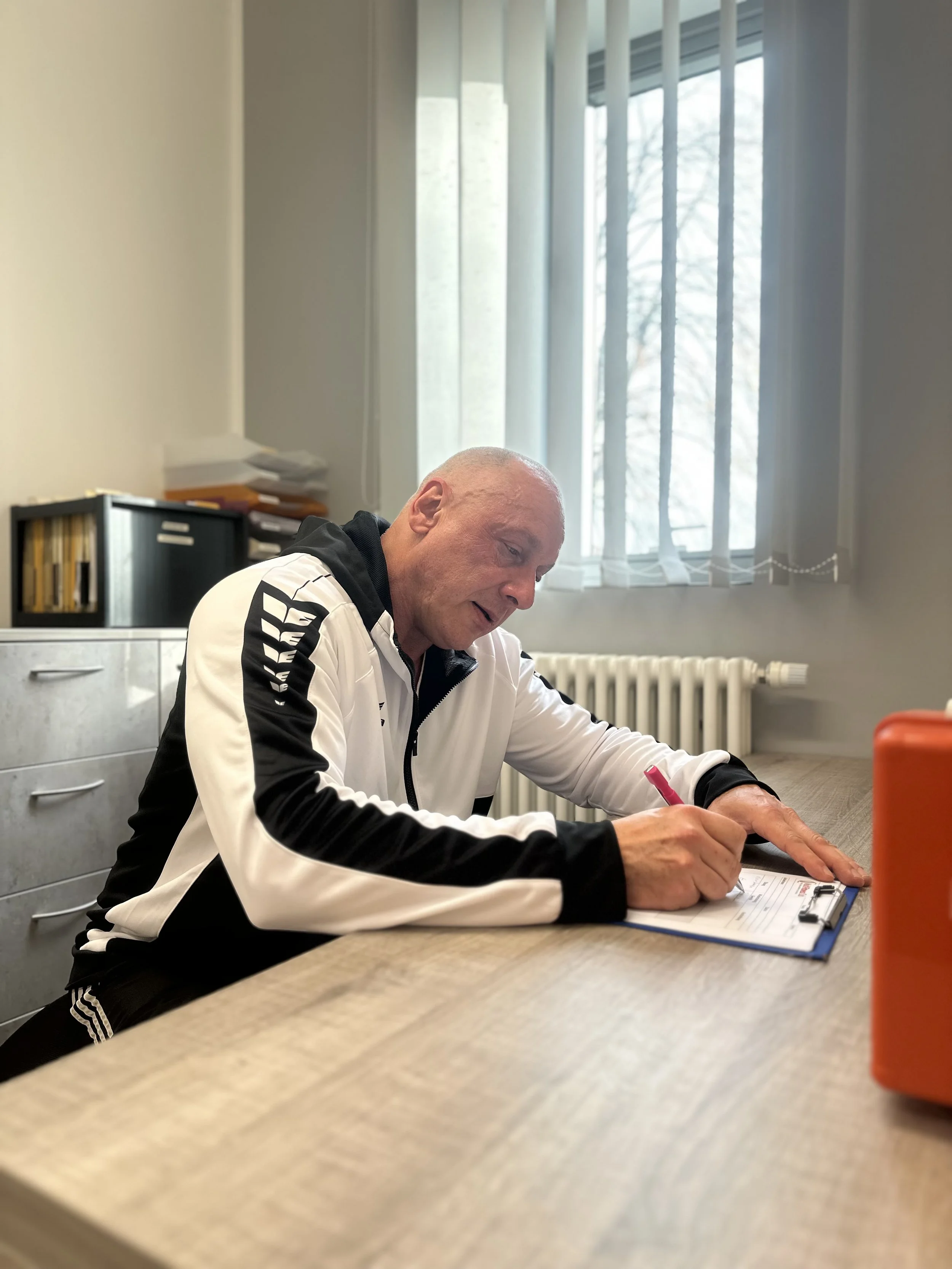 Ein Mann schreibt auf einer blauen Klarsichthülle mit einem roten Stift an einem Tisch in einem Büro, im Hintergrund sind Fenster mit weißen Vorhängen und ein Heizkörper