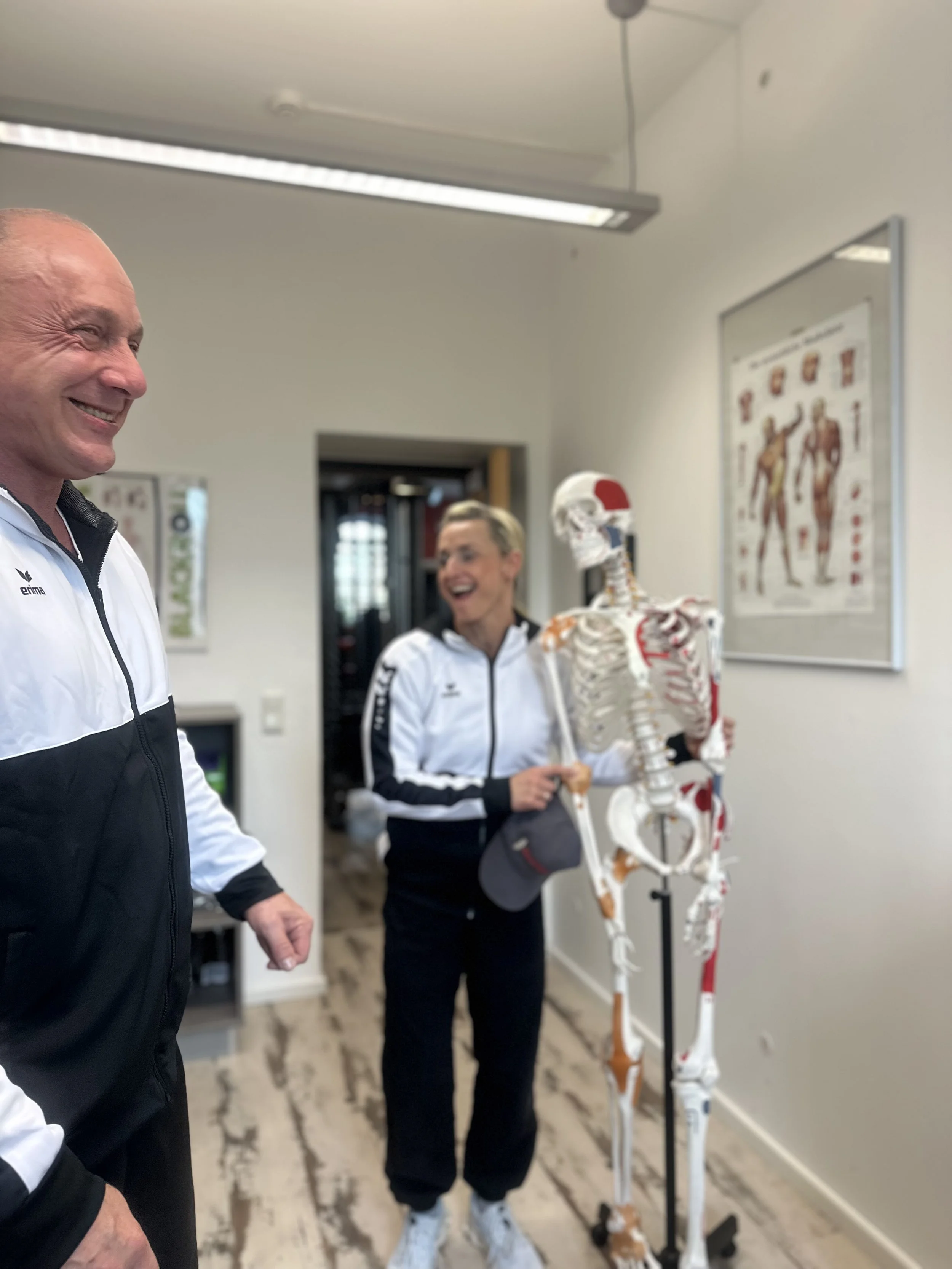 Zwei lachende Frauen und ein lachender Mann stehen in einem Raum mit einem menschlichen Skelettmodell. Die Frauen tragen weiße Sportjacken, der Mann trägt eine schwarze und weiße Jacke.