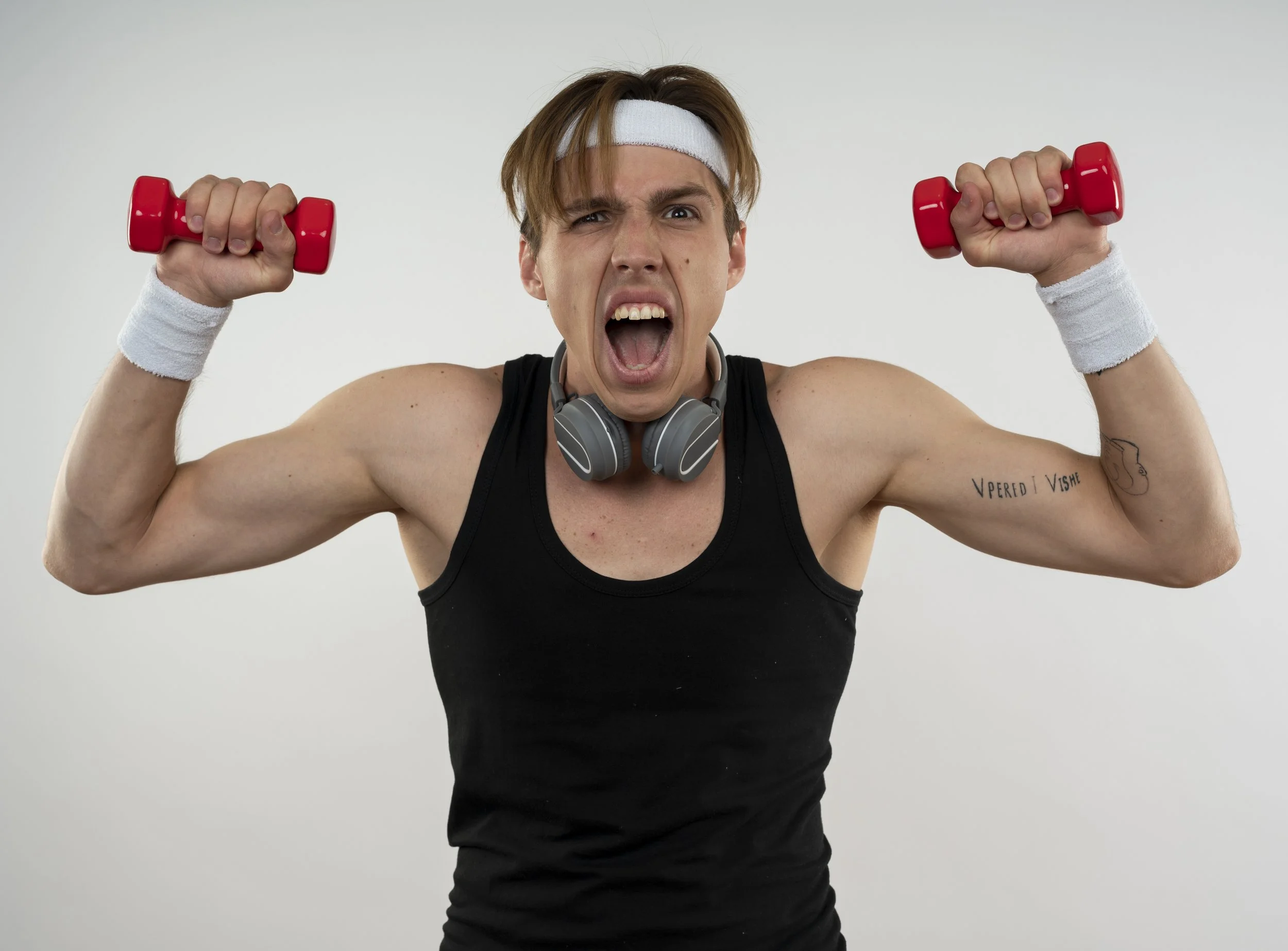 Junge Person in schwarzen Tanktop mit Kopfhörer um den Hals, weiße Kopfband, weiße Handgelenkbänder, hält kleine rote Hanteln, schreit, gegen weißen Hintergrund.