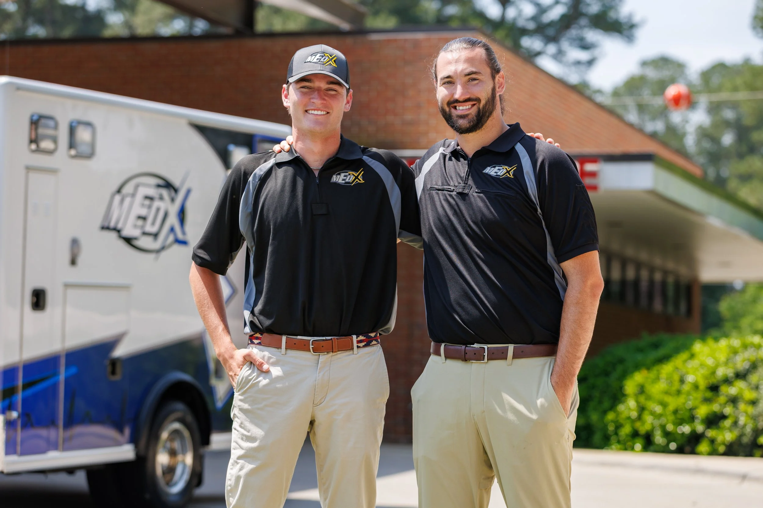 Two men standing outdoors, smiling, wearing black polo shirts with a logo, with a mobile service truck in the background, and greenery around.