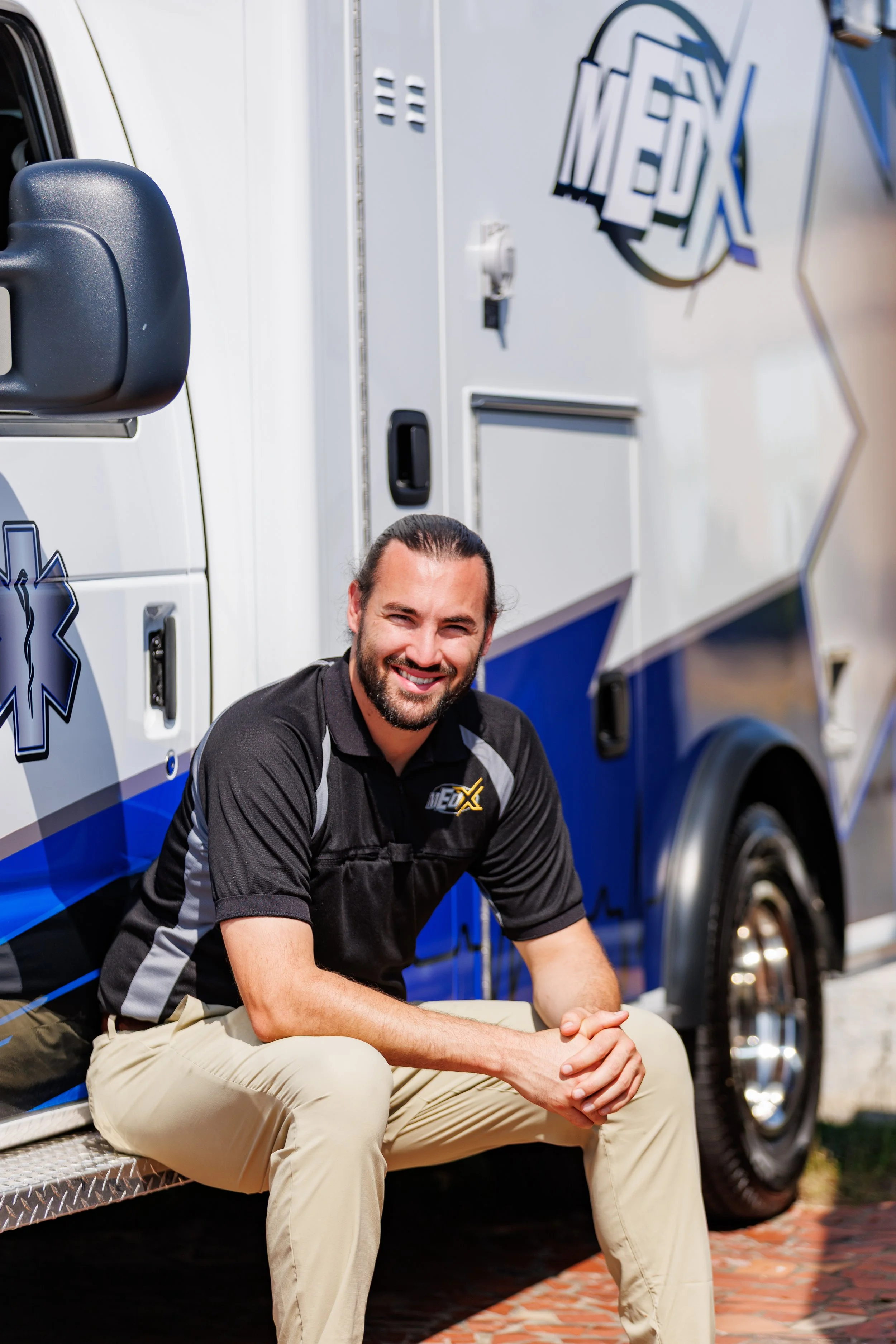 A smiling man with dark hair and a beard, wearing a black and gray EMS shirt and khaki pants, sitting on a step near an ambulance with EMS logos.