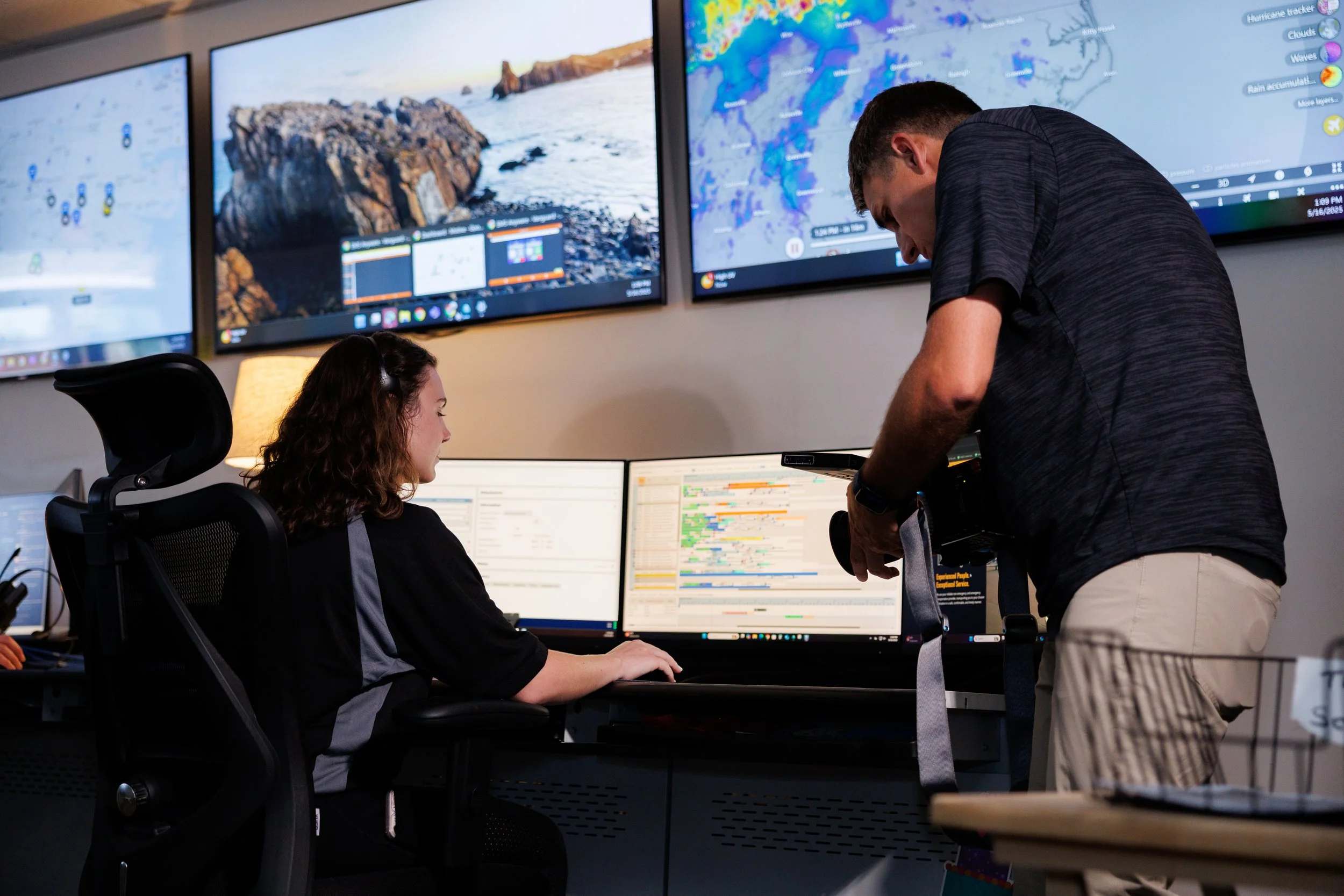 Two people working at computer stations in a control room with multiple large screens displaying weather maps and data.