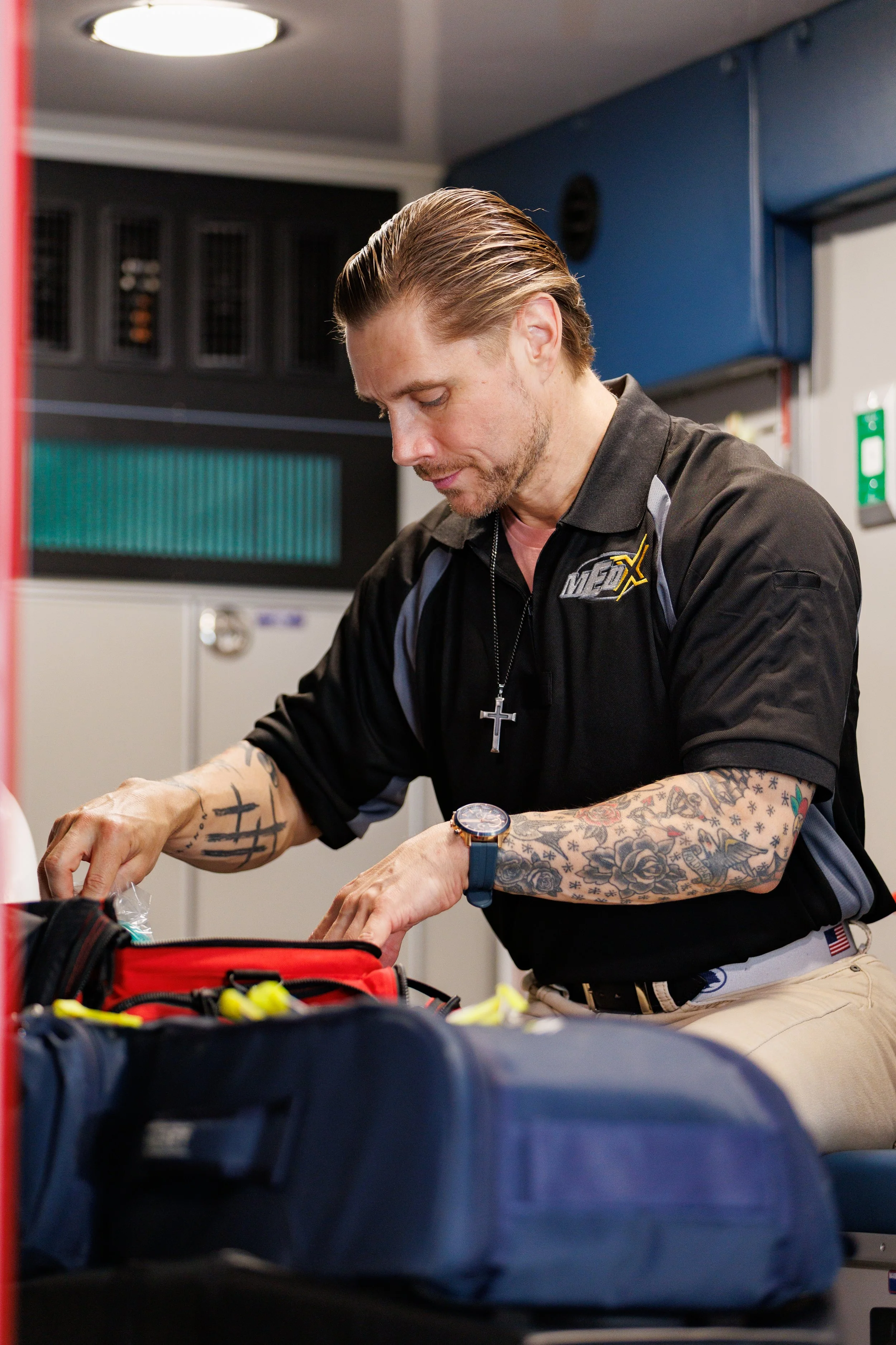 A man with tattoos on his arms, wearing a black polo shirt with a logo, a cross necklace, and a watch, is organizing emergency medical supplies on a table in an indoor setting.