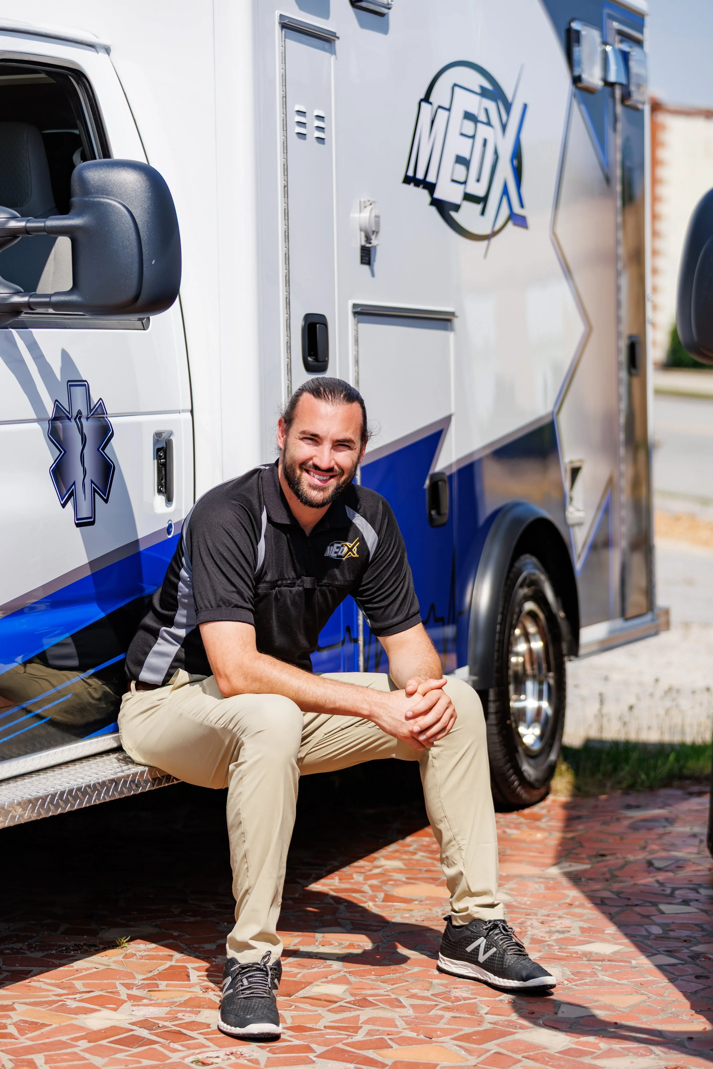 A smiling man sitting on the ground next to an ambulance, wearing a black and gray uniform shirt with the NEDx logo and light-colored pants.