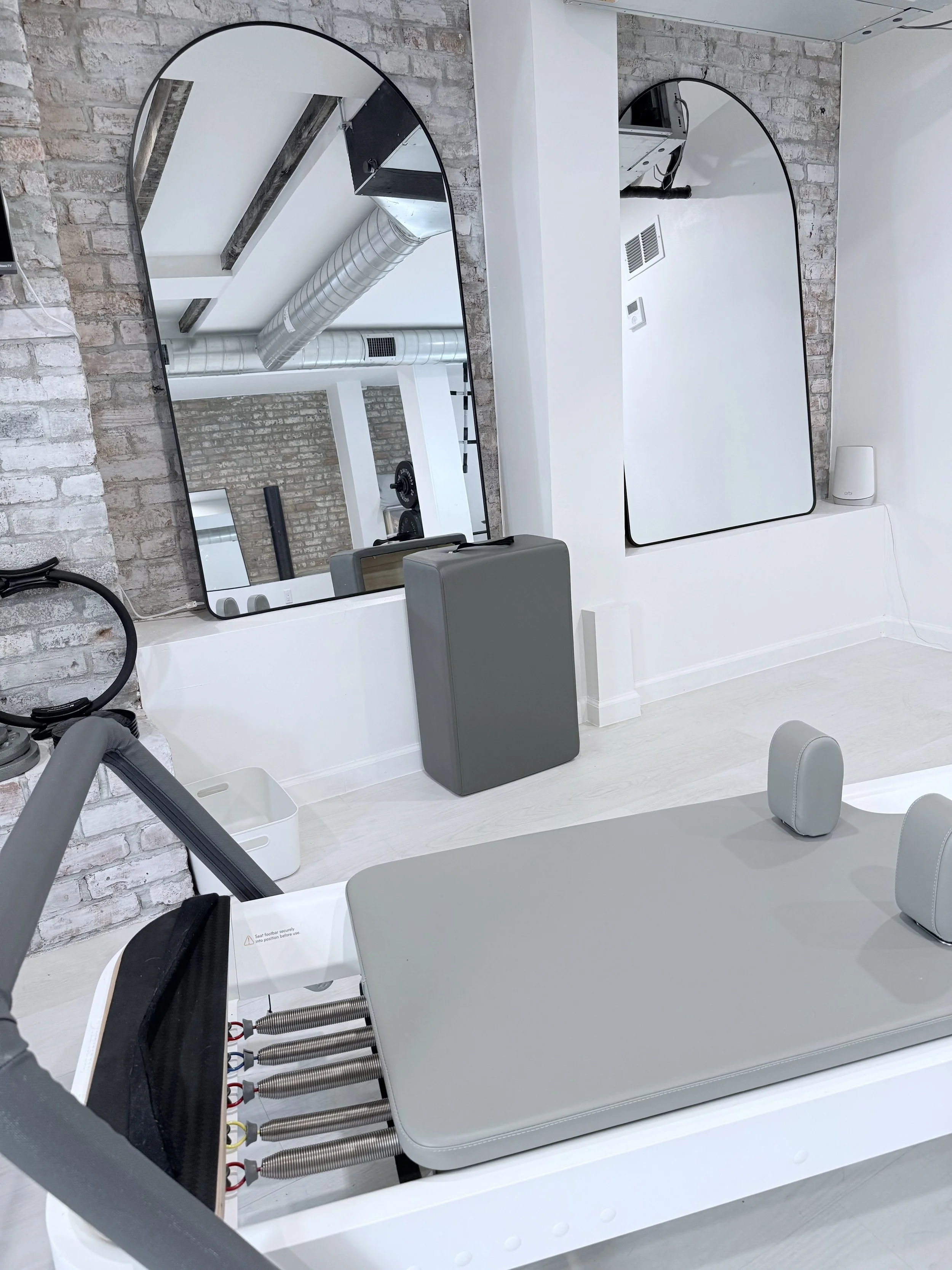 Medical examination room with a padded examination table, mirrors on white walls, exposed brick wall, visible ventilation duct, and medical equipment.